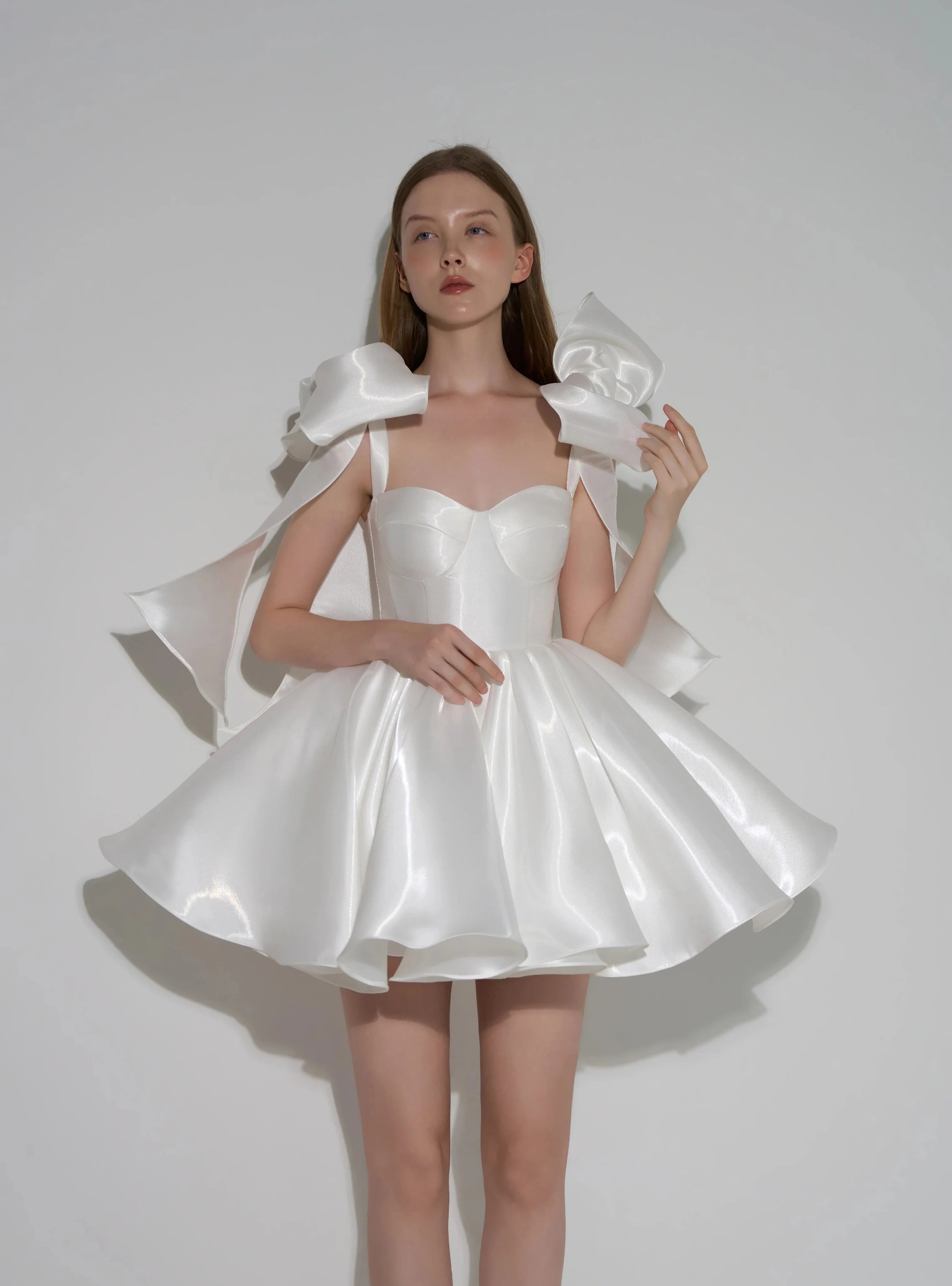 A model wearing a white satin formal dress with dramatic oversized shoulder bows and a flared skirt, posed against a plain light gray studio background. Text at the bottom reads 'Customized formal dress'.