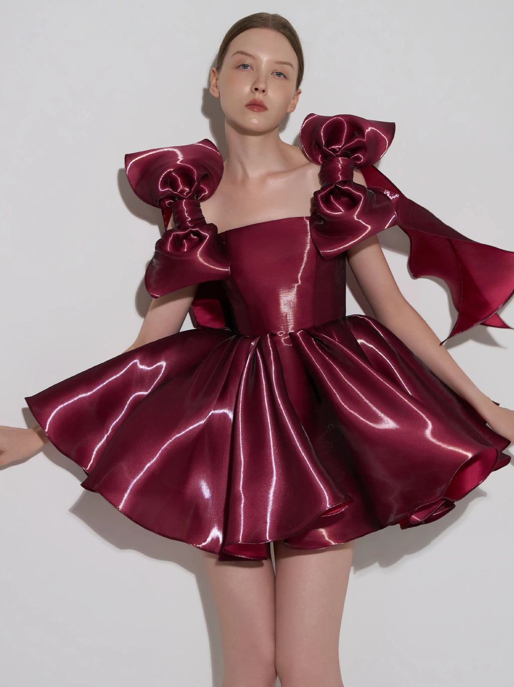 A model wearing a shiny burgundy satin formal dress with oversized bow details on the shoulders and a flared skirt, posed against a plain light gray background.