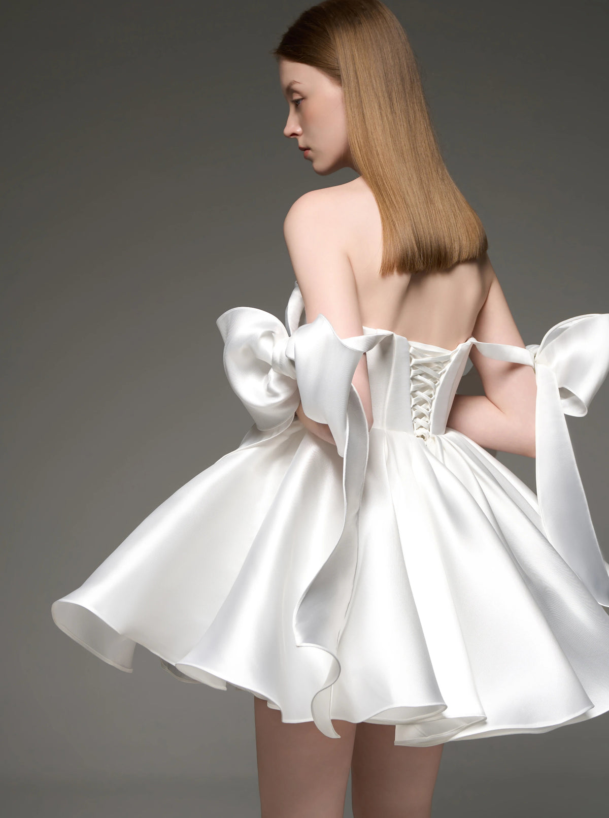 A model shown from the back wearing a white satin off-the-shoulder dress with a structured corset back and large bow details, photographed against a dark gray studio background.