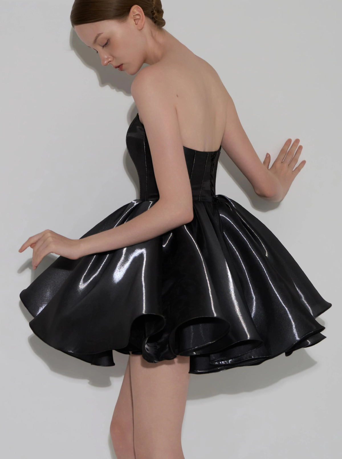 A model wearing a strapless black latex-like mini dress with a structured corset-style bodice and a flared, layered skirt, posed against a plain light gray background.
