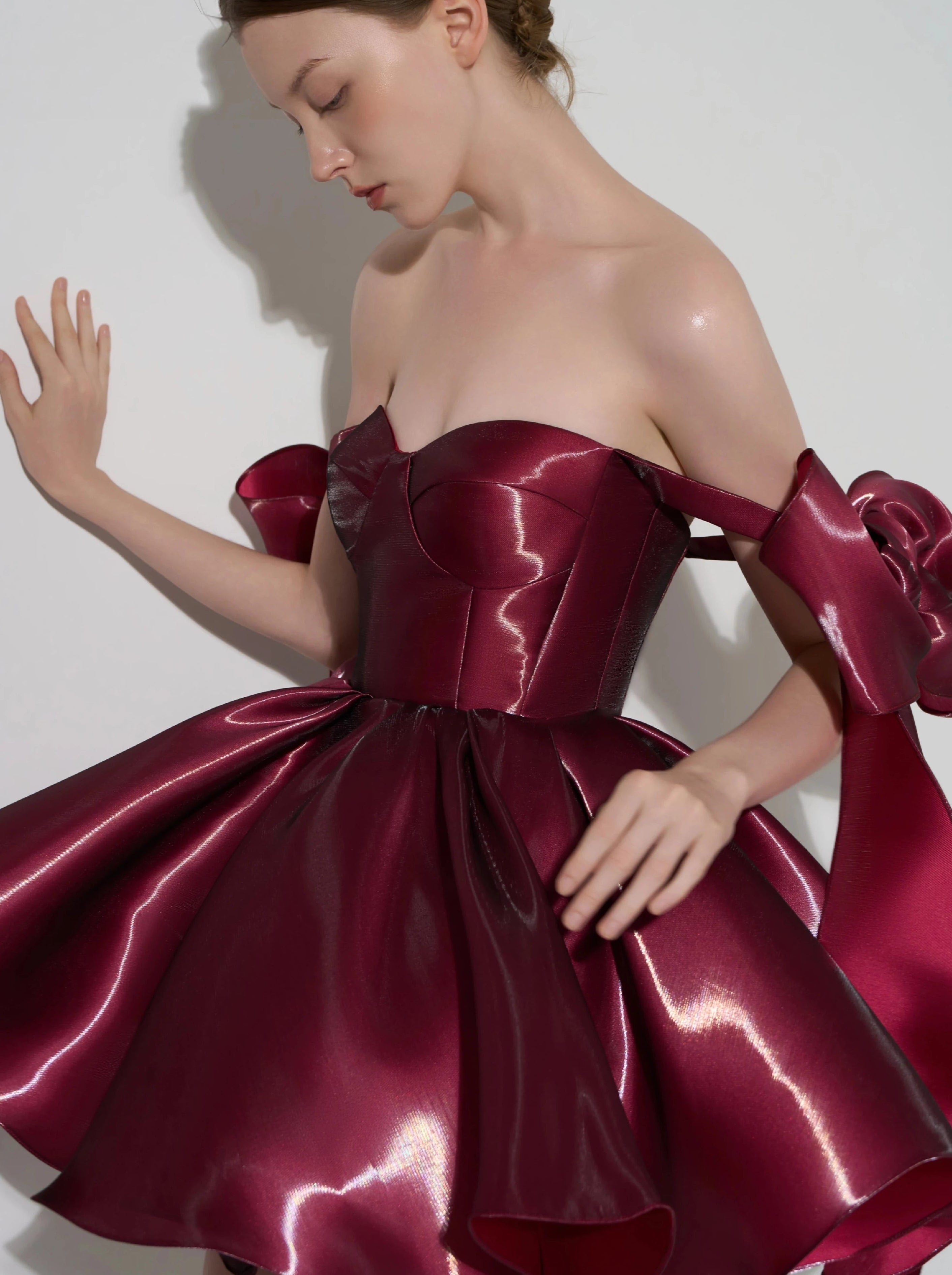 A woman modeling a strapless off-the-shoulder formal dress made of shiny deep burgundy satin, with a structured corset-style bodice and a full skirt with large bow details at the shoulders, against a plain light gray studio background.
