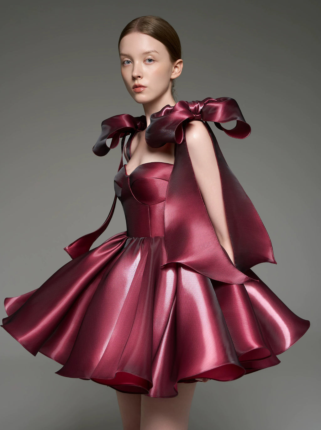 A model wearing a custom formal dress made of shiny burgundy satin, featuring a structured corset bodice, a flared skirt, and dramatic oversized bow details on the shoulders, set against a solid gray studio background.