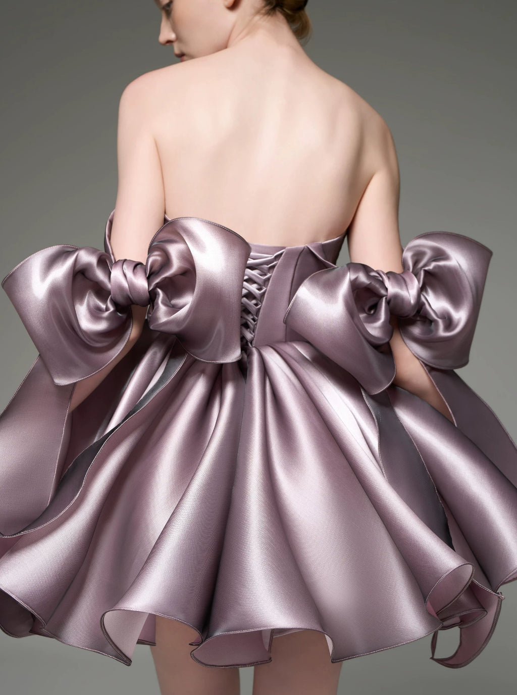 A model shown from the back wearing a strapless formal dress made of shiny lavender-purple satin, featuring oversized bow details on the shoulders and a flared skirt, against a neutral gray studio background.