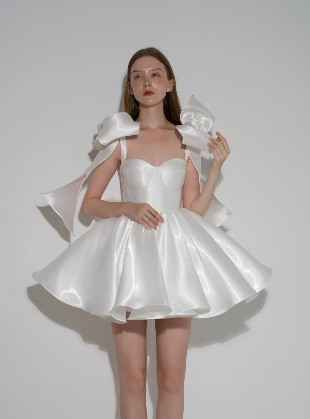 A model wearing a white satin formal dress with dramatic oversized shoulder bows and a flared skirt, posed against a plain light gray studio background. Text at the bottom reads 'Customized formal dress'.