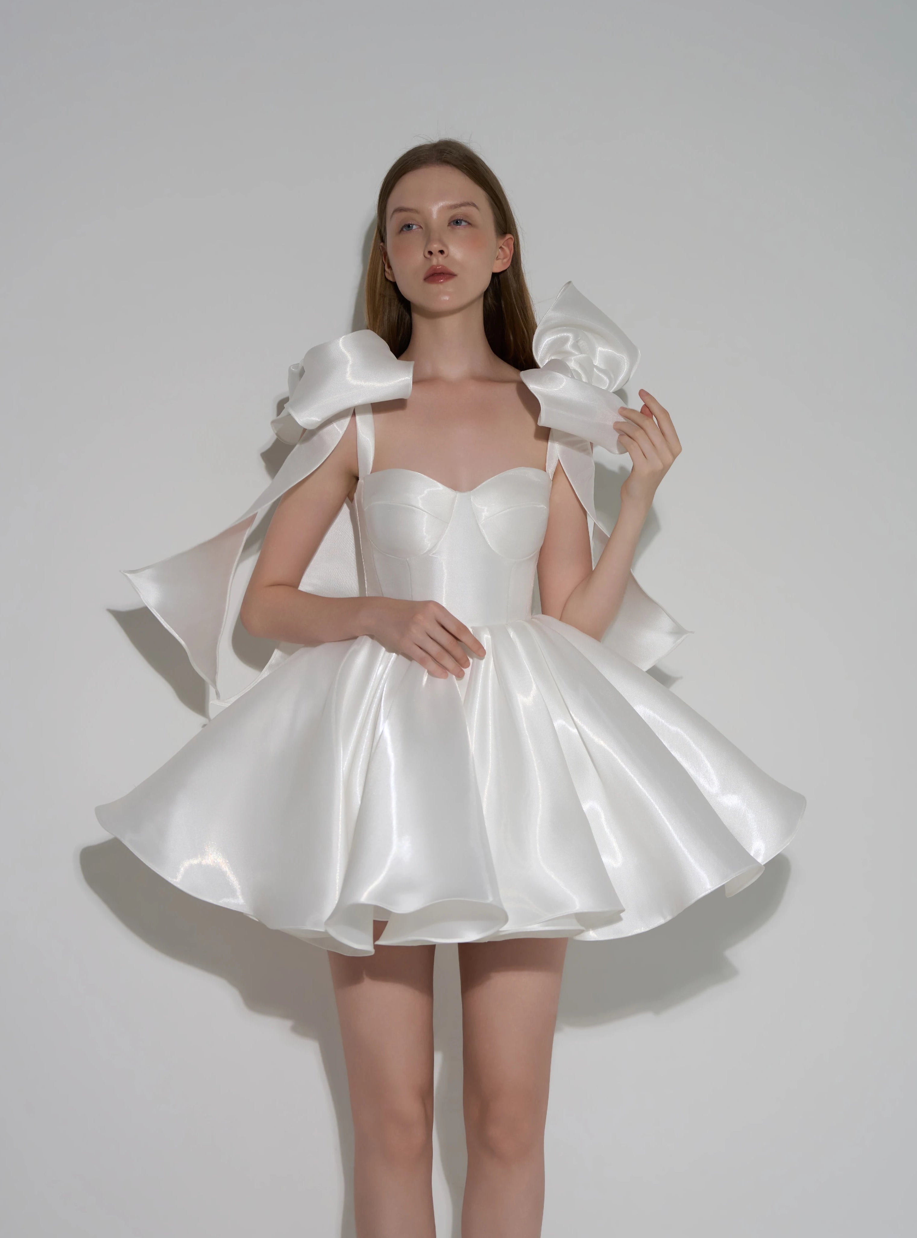 A model wearing a white satin formal dress with dramatic oversized shoulder bows and a flared skirt, posed against a plain light gray studio background. Text at the bottom reads 'Customized formal dress'.
