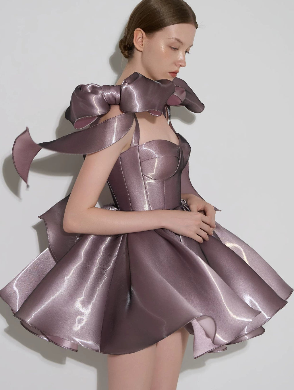 A model wearing a metallic mauve mini dress with oversized bow details on the shoulders and a structured corset bodice, set against a plain light gray background.