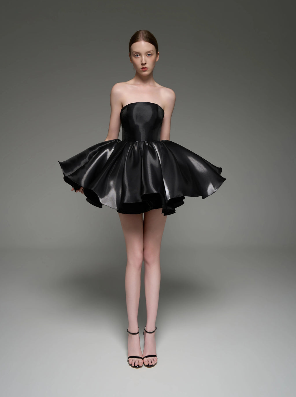 A model wearing a black strapless mini dress with a flared, tiered skirt, standing against a dark gray studio background. The brand name 'Lusilk' is displayed at the top of the image.