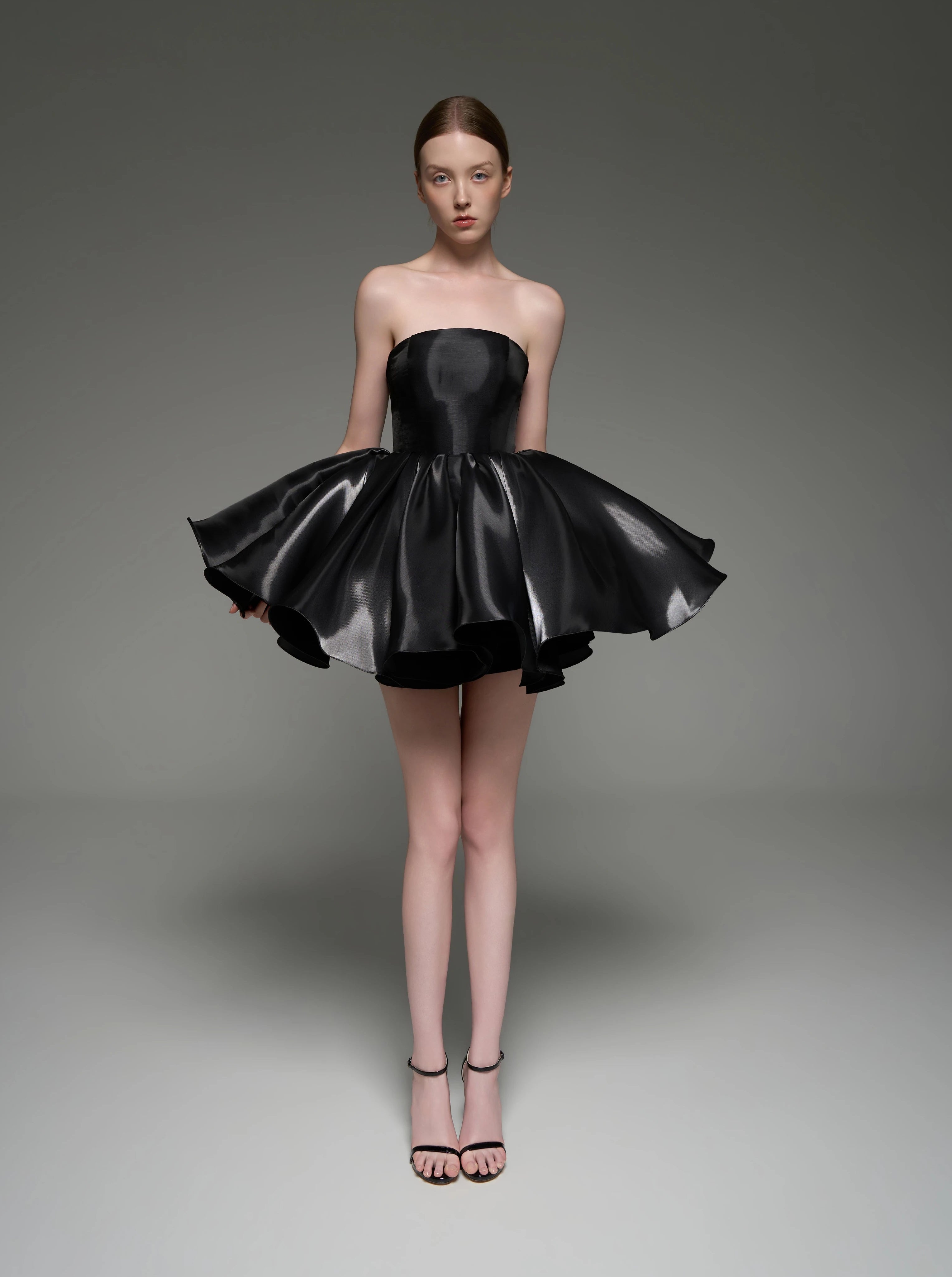 A model wearing a black strapless mini dress with a flared, tiered skirt, standing against a dark gray studio background. The brand name 'Lusilk' is displayed at the top of the image.