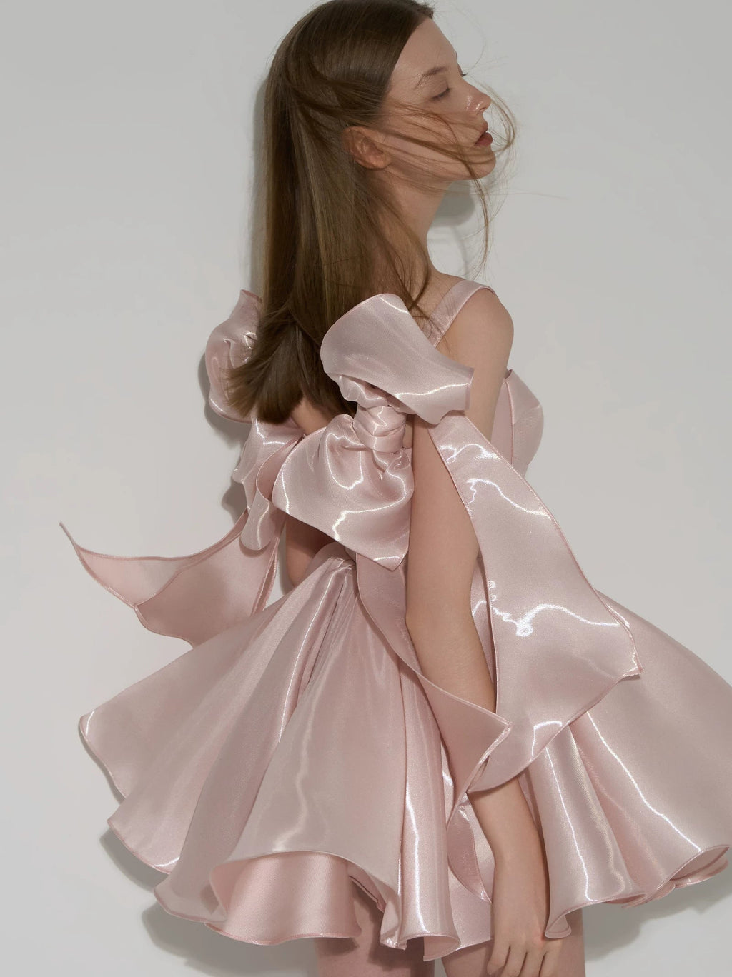 A woman wearing a shiny pale pink satin dress with dramatic oversized bow details and layered ruffled sleeves, photographed against a plain light gray studio background.