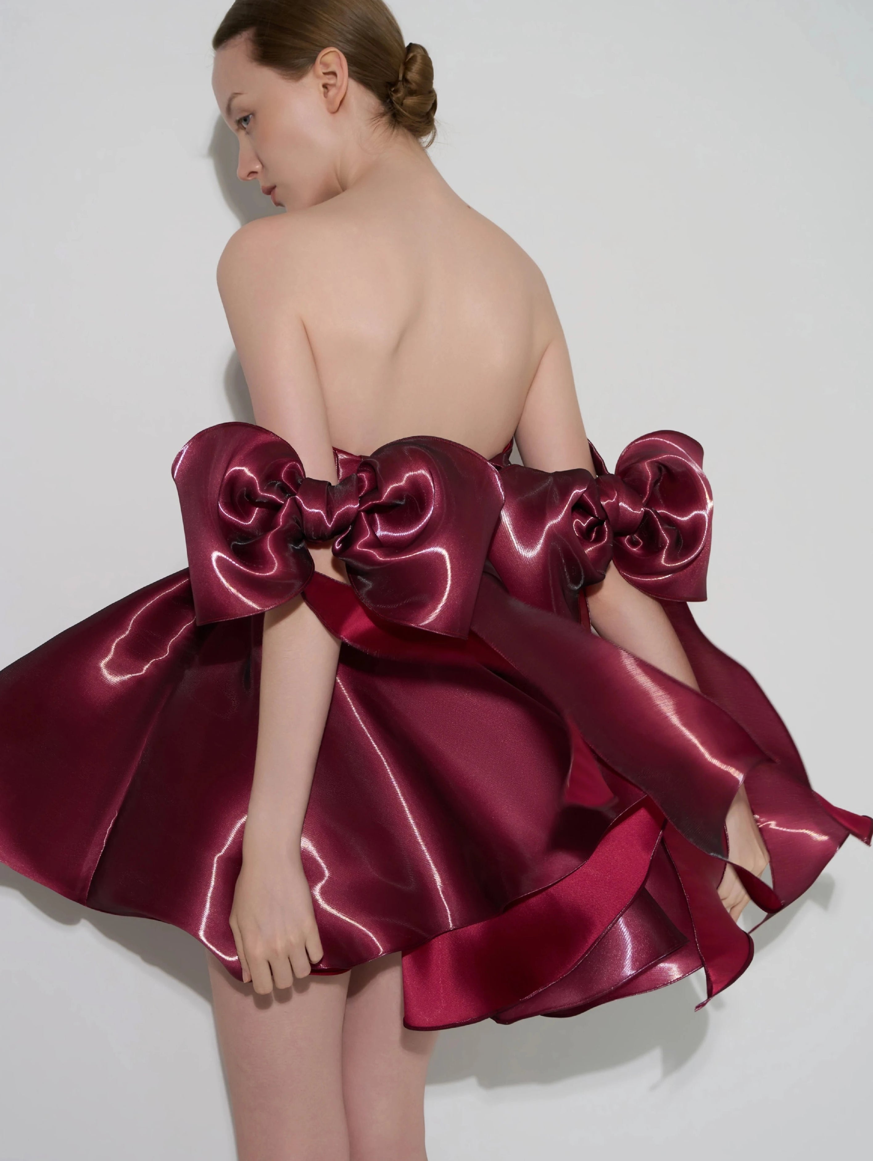 A model shown from the back wearing a strapless formal dress with oversized bow details and a voluminous draped skirt in deep burgundy satin against a light gray studio background.