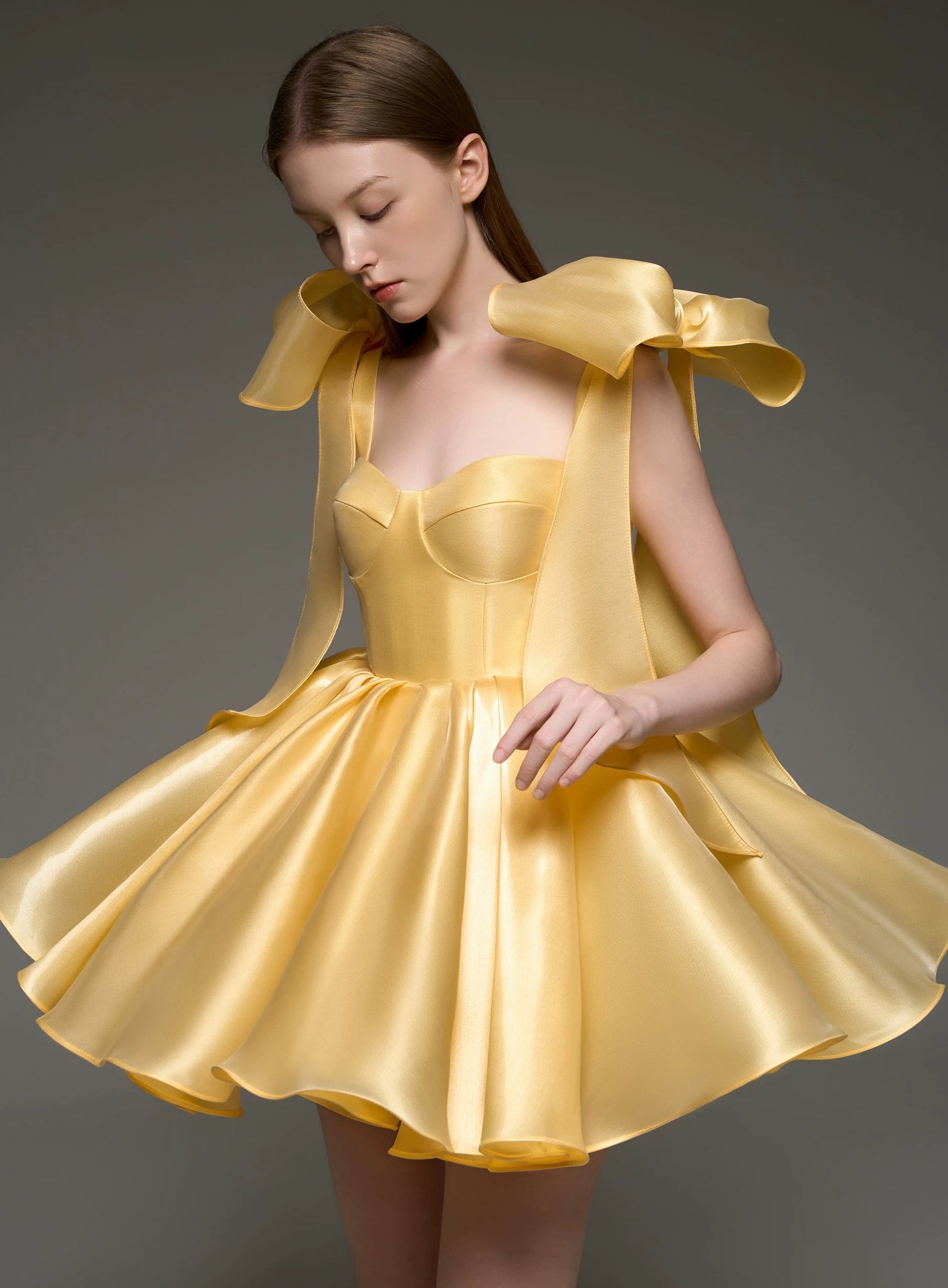 A woman modeling a short formal dress made of shiny satin-like yellow fabric, with oversized bow details on the shoulders and a flared skirt, photographed against a dark gray studio background.