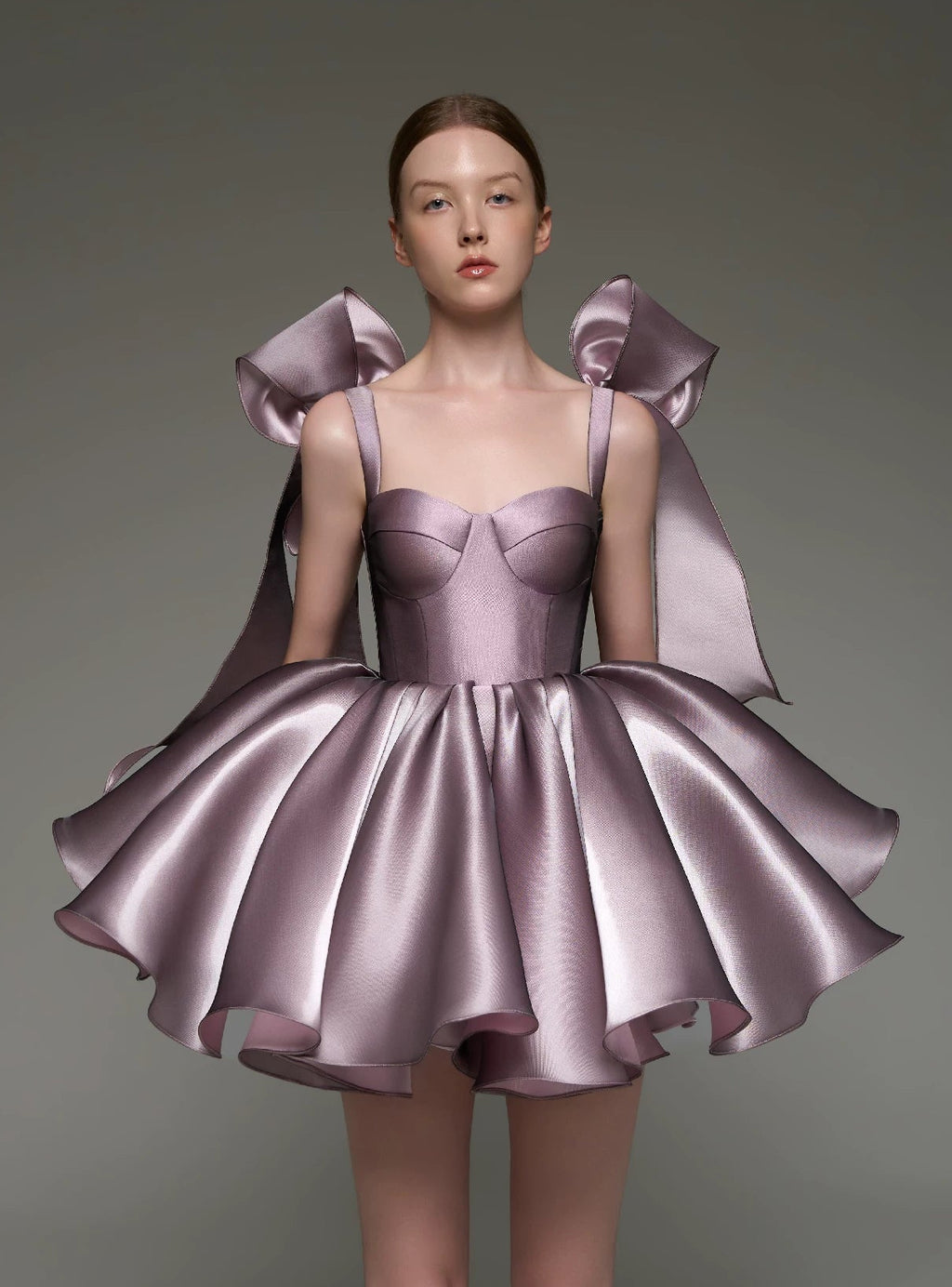 A model wearing a lavender satin mini dress with structured shoulder bows, photographed against a neutral gray studio background.