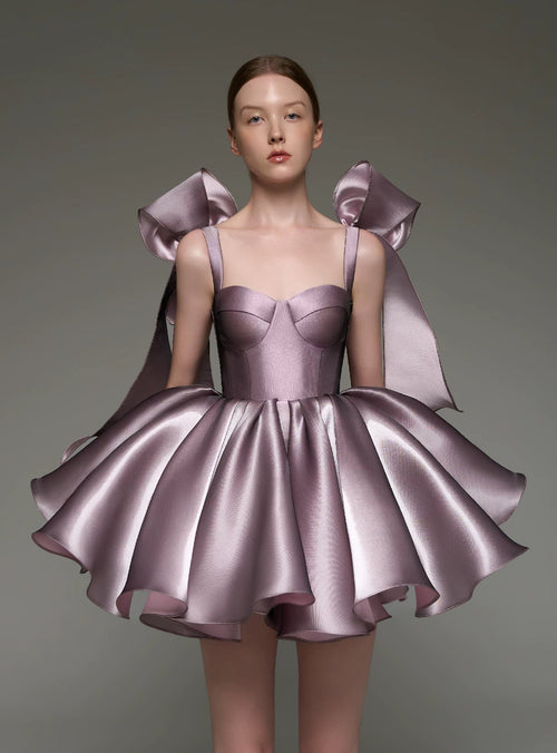A model wearing a lavender satin mini dress with structured shoulder bows, photographed against a neutral gray studio background.