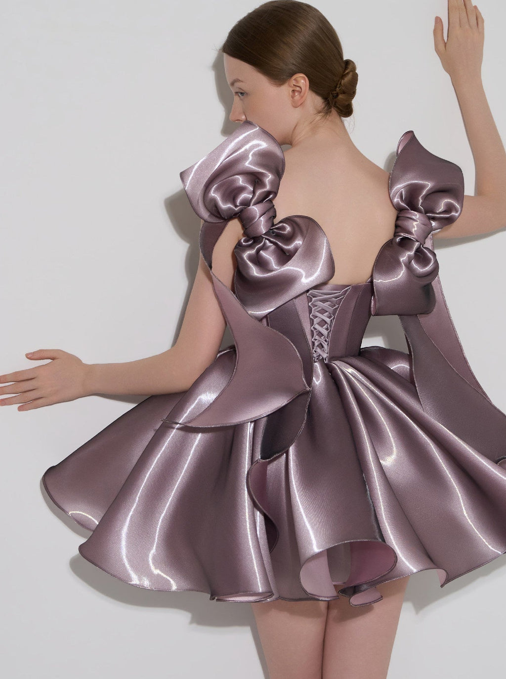 A model shown from the back wearing a short, shiny mauve-purple satin dress with oversized bow details on the shoulders and a lace-up corset back, against a plain light gray studio background.
