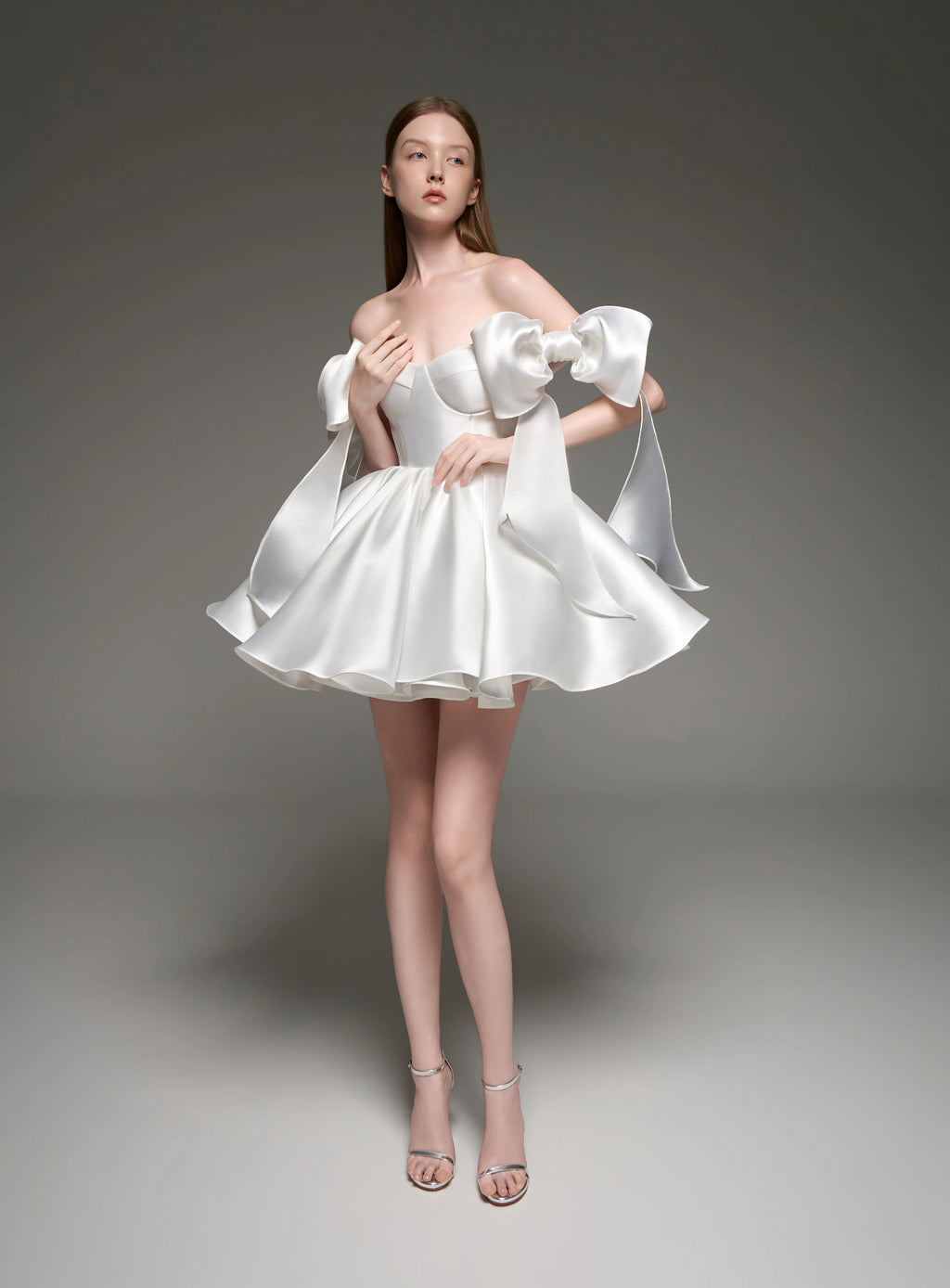 A model wearing a white satin off-the-shoulder mini dress with dramatic puffed sleeves and a flared skirt, standing against a dark gray studio background. The brand name 'Lusilk' is displayed at the top.