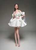 A model wearing a white satin off-the-shoulder mini dress with dramatic puffed sleeves and a flared skirt, standing against a dark gray studio background. The brand name 'Lusilk' is displayed at the top.