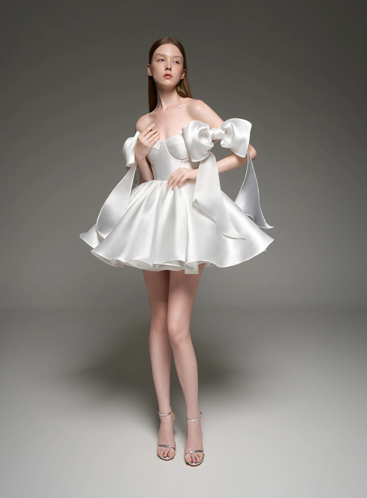 A model wearing a white satin off-the-shoulder mini dress with dramatic puffed sleeves and a flared skirt, standing against a dark gray studio background. The brand name 'Lusilk' is displayed at the top.