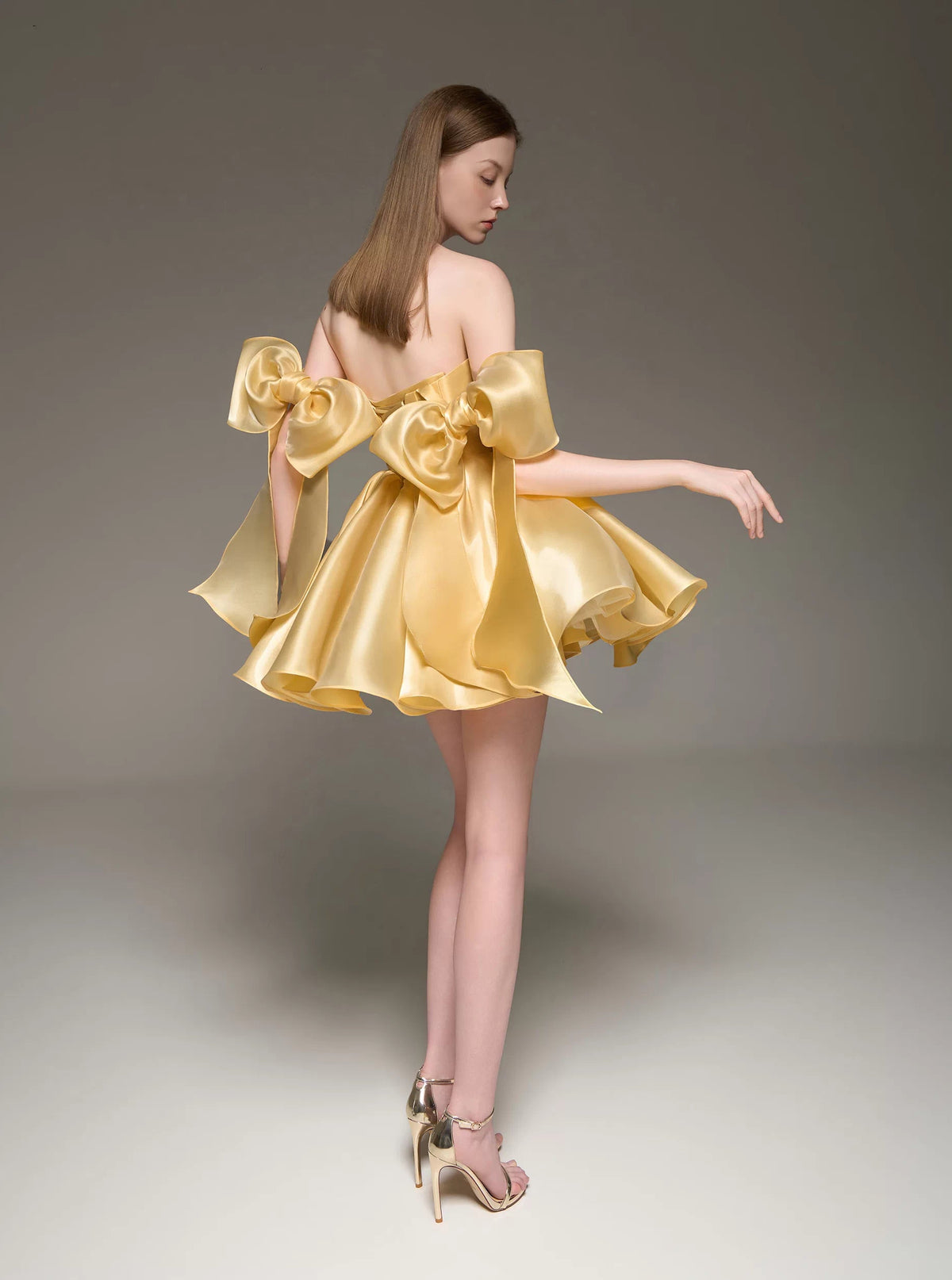 A woman modeling a short, strapless formal dress made of shiny gold satin, featuring oversized bow details on the shoulders and a flared skirt, photographed against a neutral gray studio background.