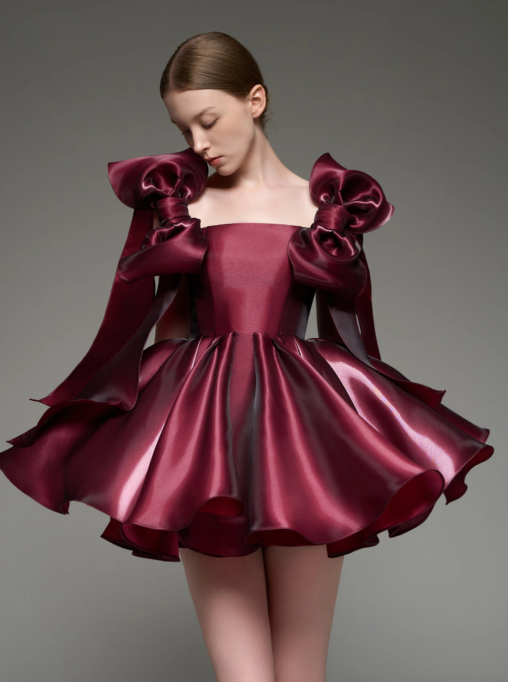 A model wearing a custom formal dress made of shiny burgundy satin, featuring oversized bow details on the shoulders and a flared skirt, set against a solid gray studio background.