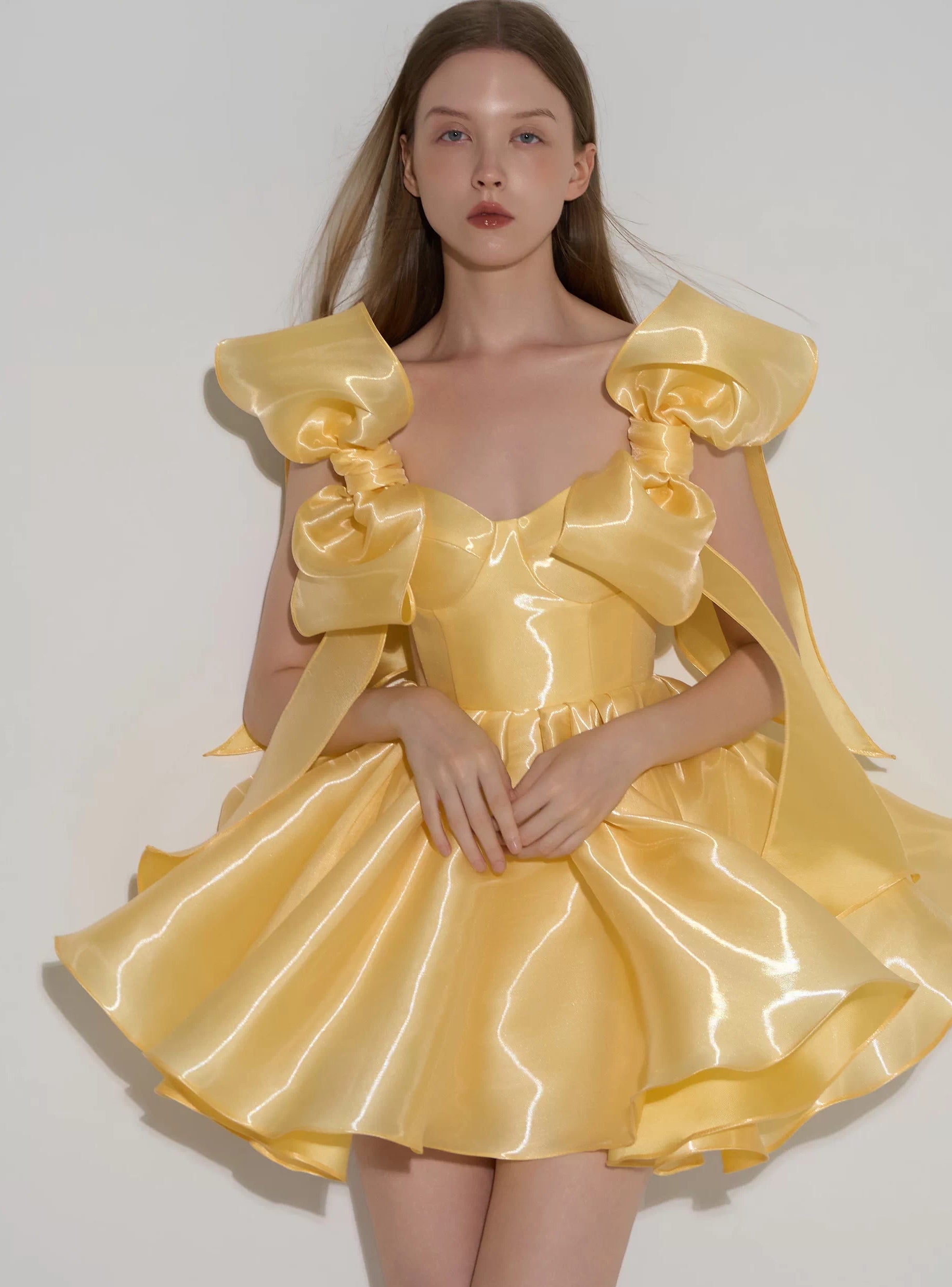 A model wearing a shiny yellow formal dress with oversized bow details on the shoulders and a flared skirt, photographed against a plain light gray background.