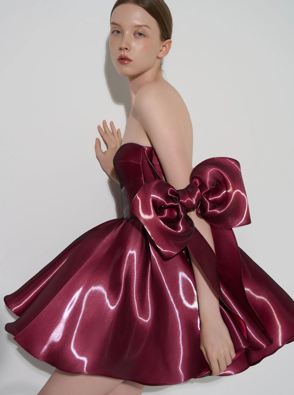 A model wearing a strapless formal dress made of shiny burgundy satin, featuring a large oversized bow at the back, posed against a light gray studio background.