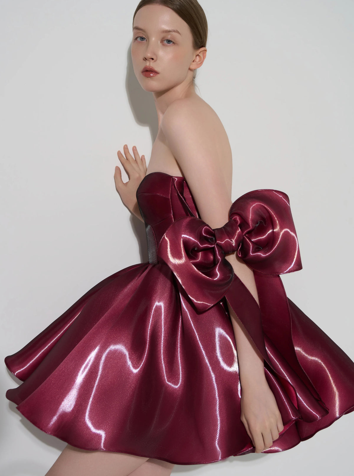 A model wearing a strapless formal dress made of shiny burgundy satin, featuring a large oversized bow at the back, posed against a light gray studio background.