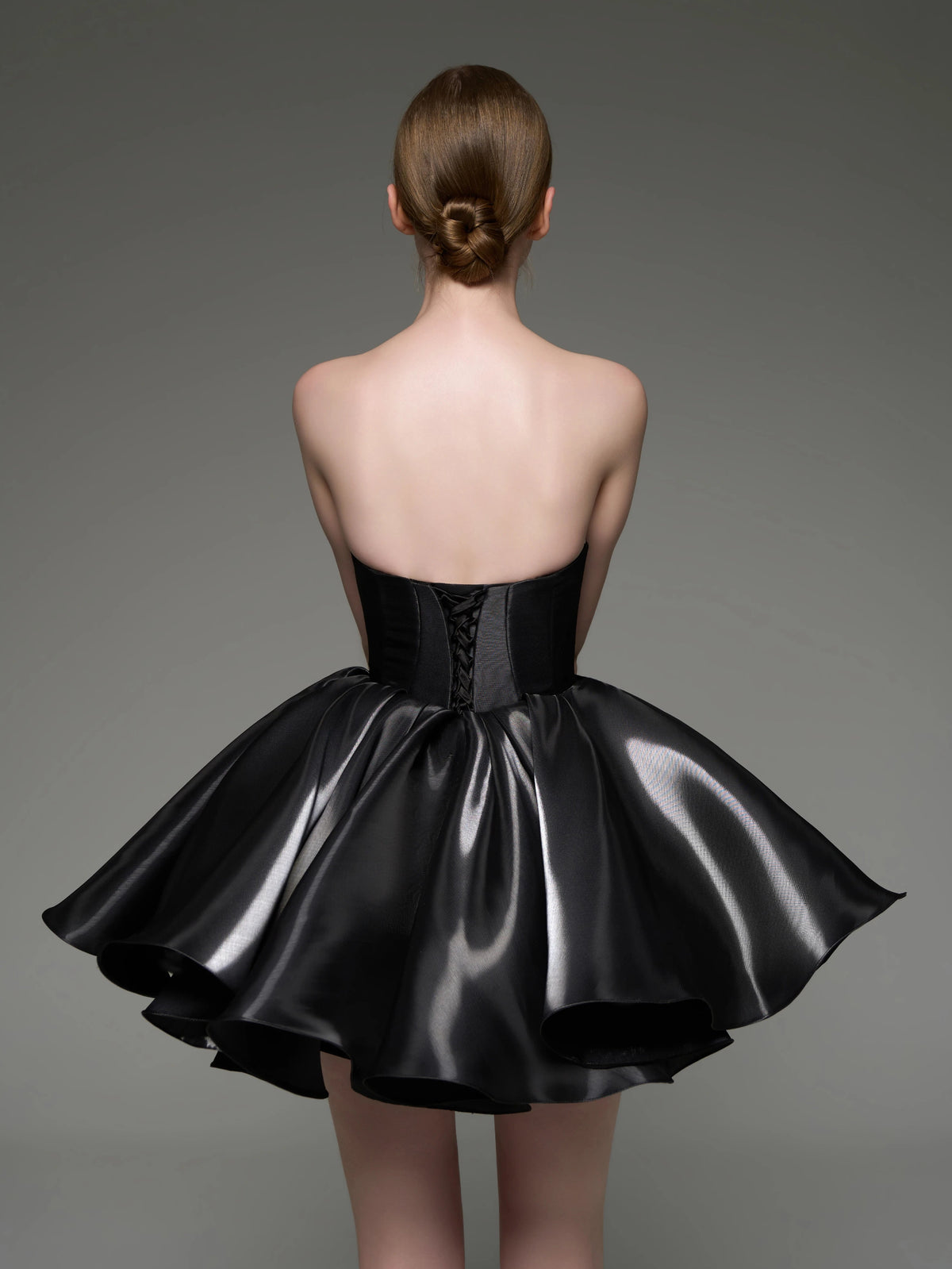 A woman shown from the back wearing a strapless black formal dress with a structured corset-style waist and a full shiny skirt, photographed against a plain gray studio background.