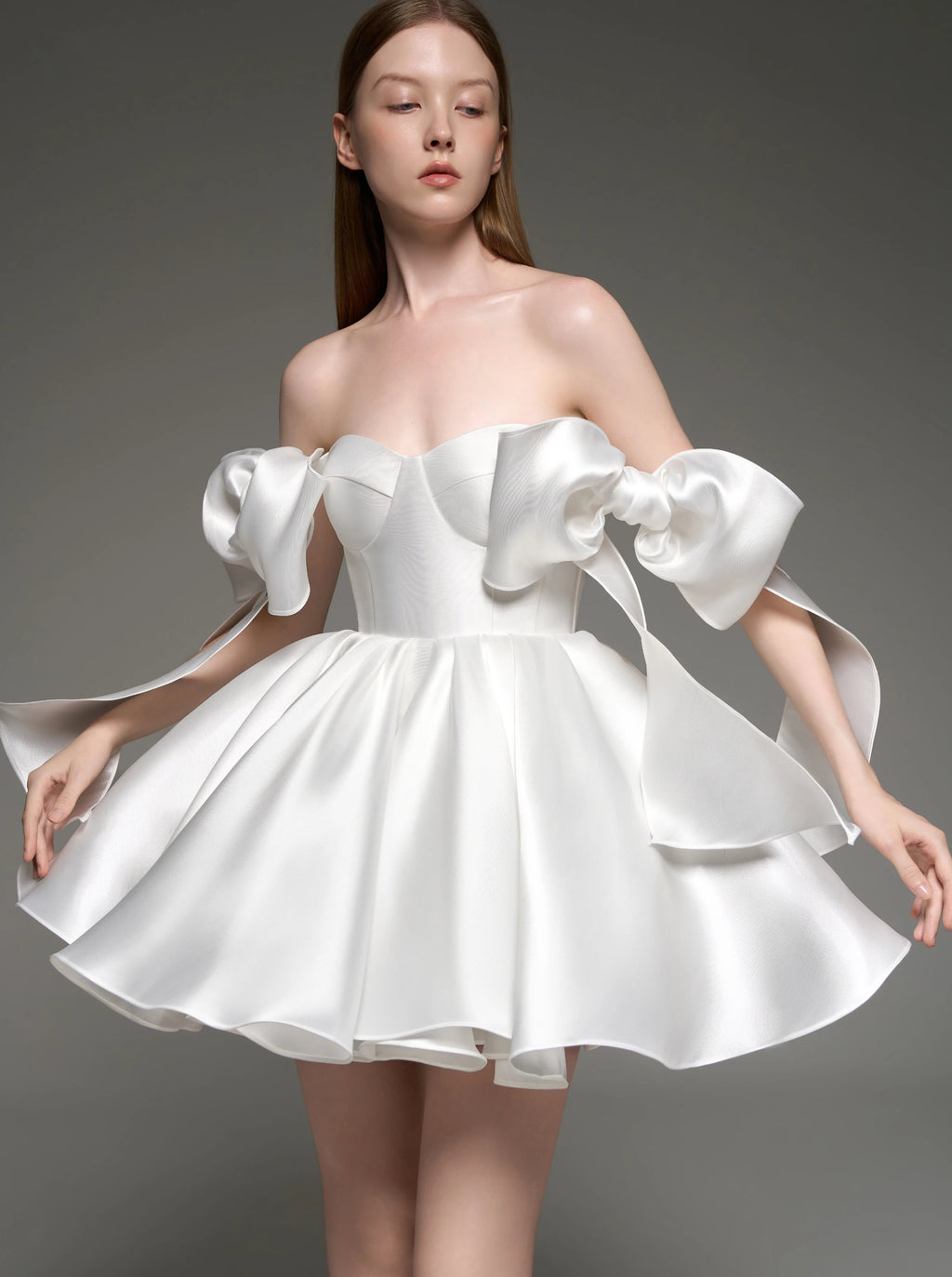 A woman modeling a white off-the-shoulder satin mini dress with dramatic puffed sleeves and a flared skirt, photographed against a dark gray studio background.