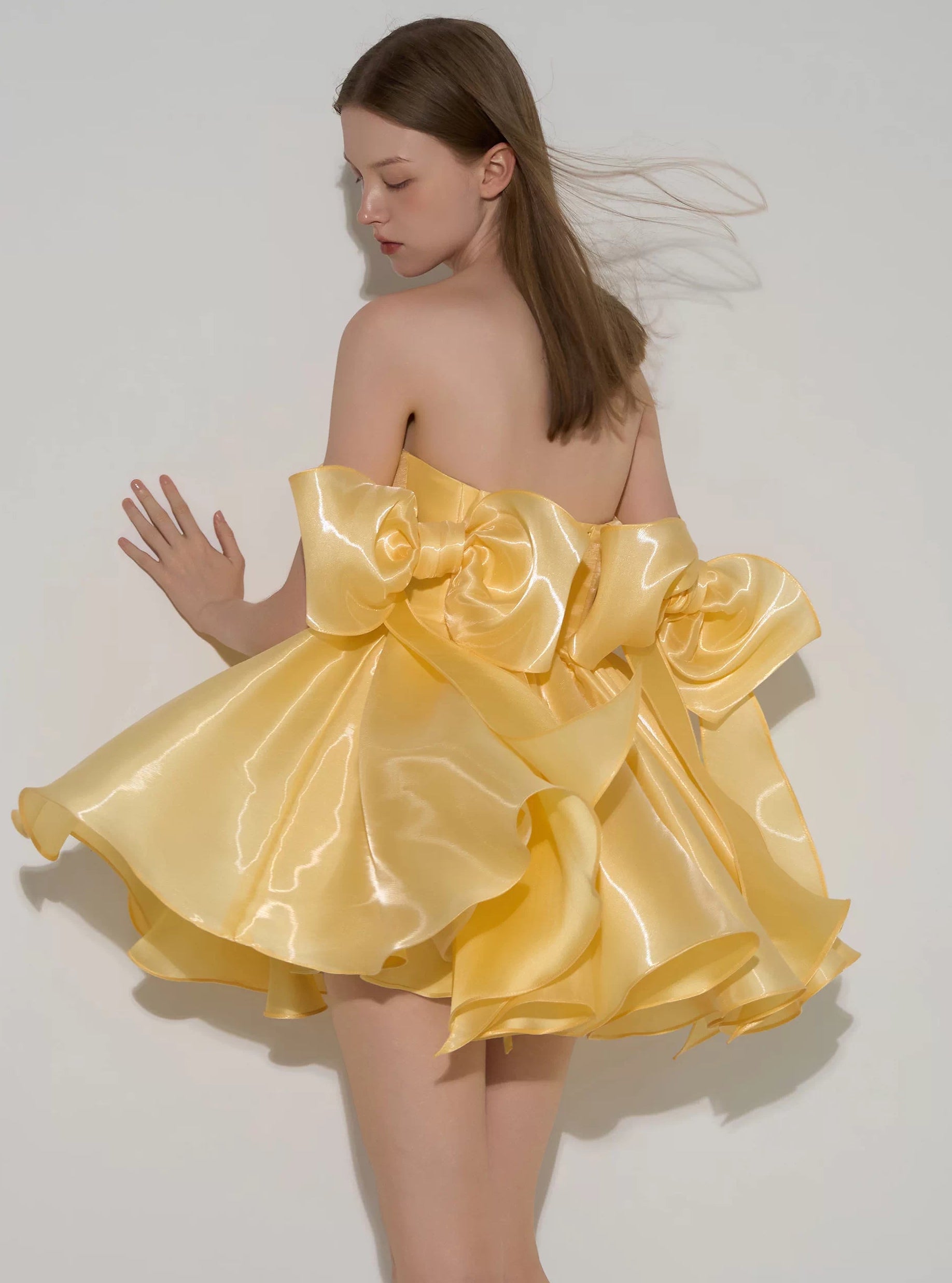 A woman modeling a strapless formal dress made of shiny yellow satin, photographed against a plain light gray studio background. The dress has dramatic oversized bow details on the back and layered ruffled fabric.