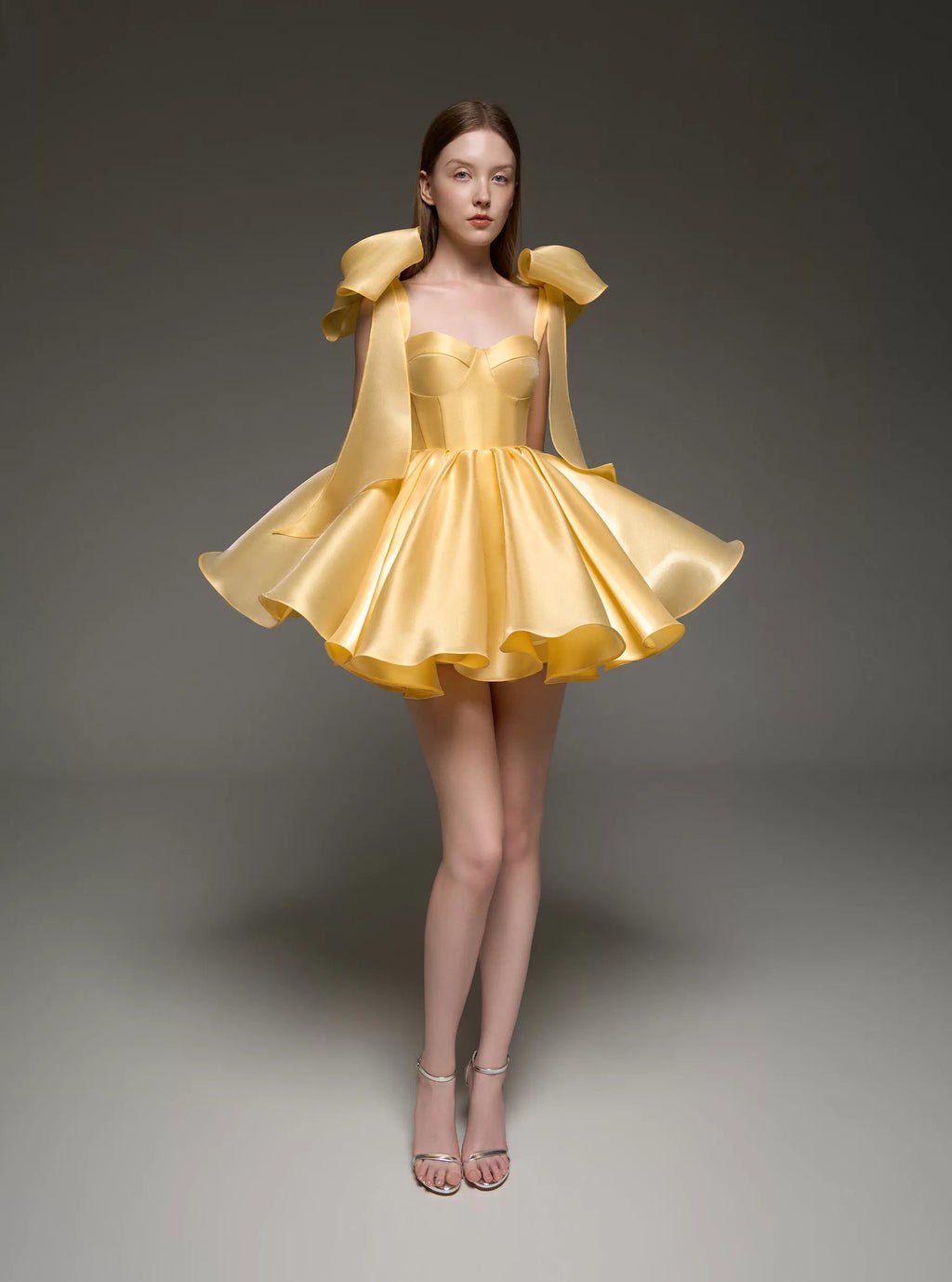 A model wearing a bright yellow formal dress with structured shoulder details and a flared skirt, standing against a dark gray studio background.