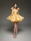 A model wearing a bright yellow formal dress with structured shoulder details and a flared skirt, standing against a dark gray studio background.
