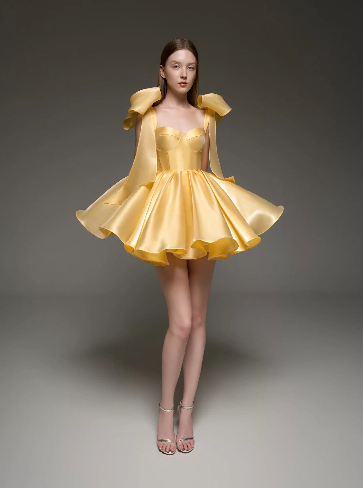 A model wearing a bright yellow formal dress with structured shoulder details and a flared skirt, standing against a dark gray studio background.