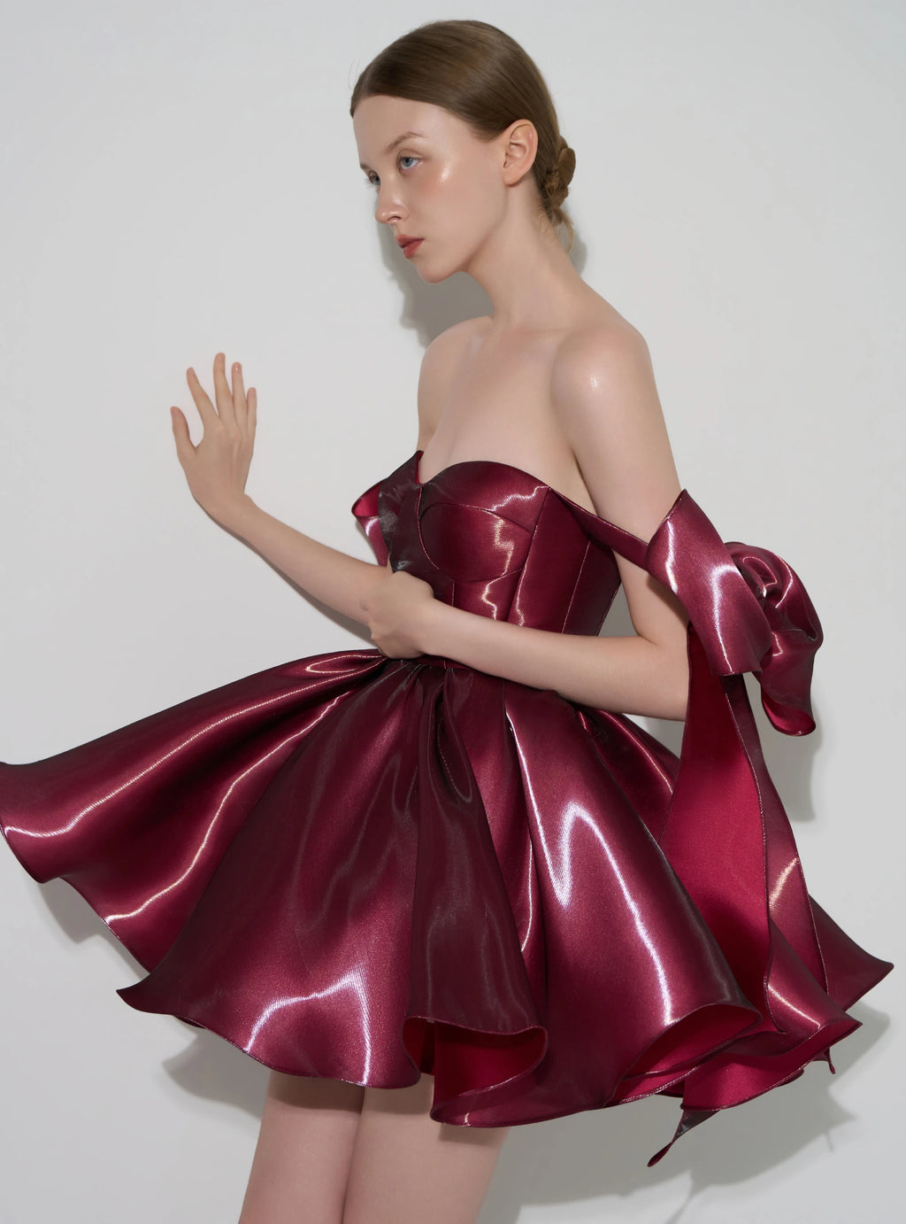 A mannequin wearing a strapless metallic burgundy mini dress with dramatic off-shoulder bow details and a flared skirt, posed against a plain light gray studio background.