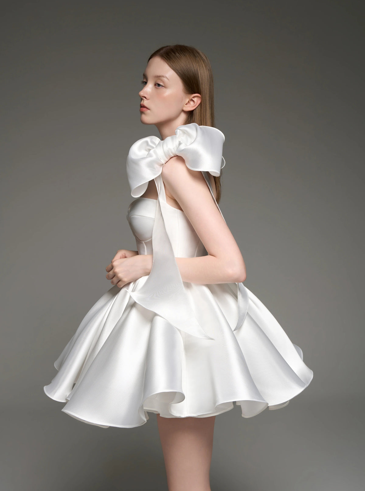 A woman modeling a white satin formal dress with a large bow at the neckline and a flared skirt, photographed against a plain gray studio background.