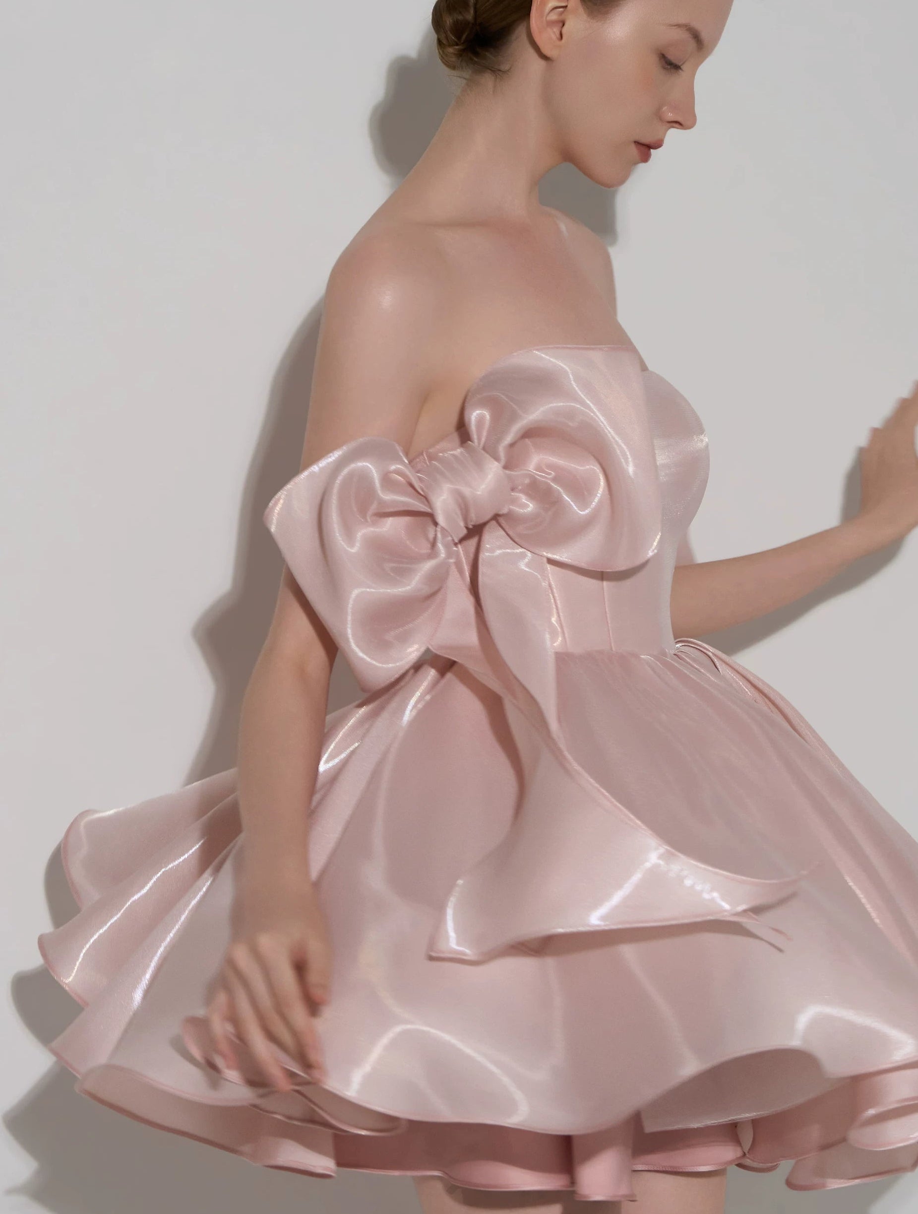 A woman wearing a strapless light pink satin dress with a large oversized bow at the bodice and a flared ruffled skirt, posed against a plain light gray studio background.