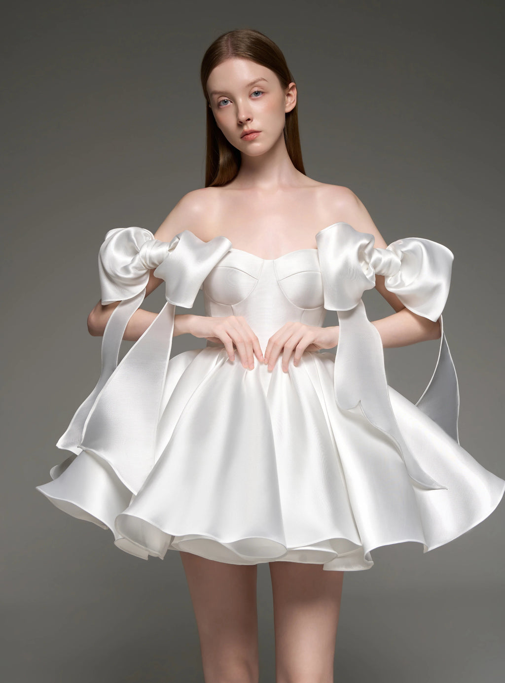 A model wearing a white satin off-the-shoulder mini dress with dramatic oversized bow details on the sleeves and a flared skirt, posed against a dark gray studio background.