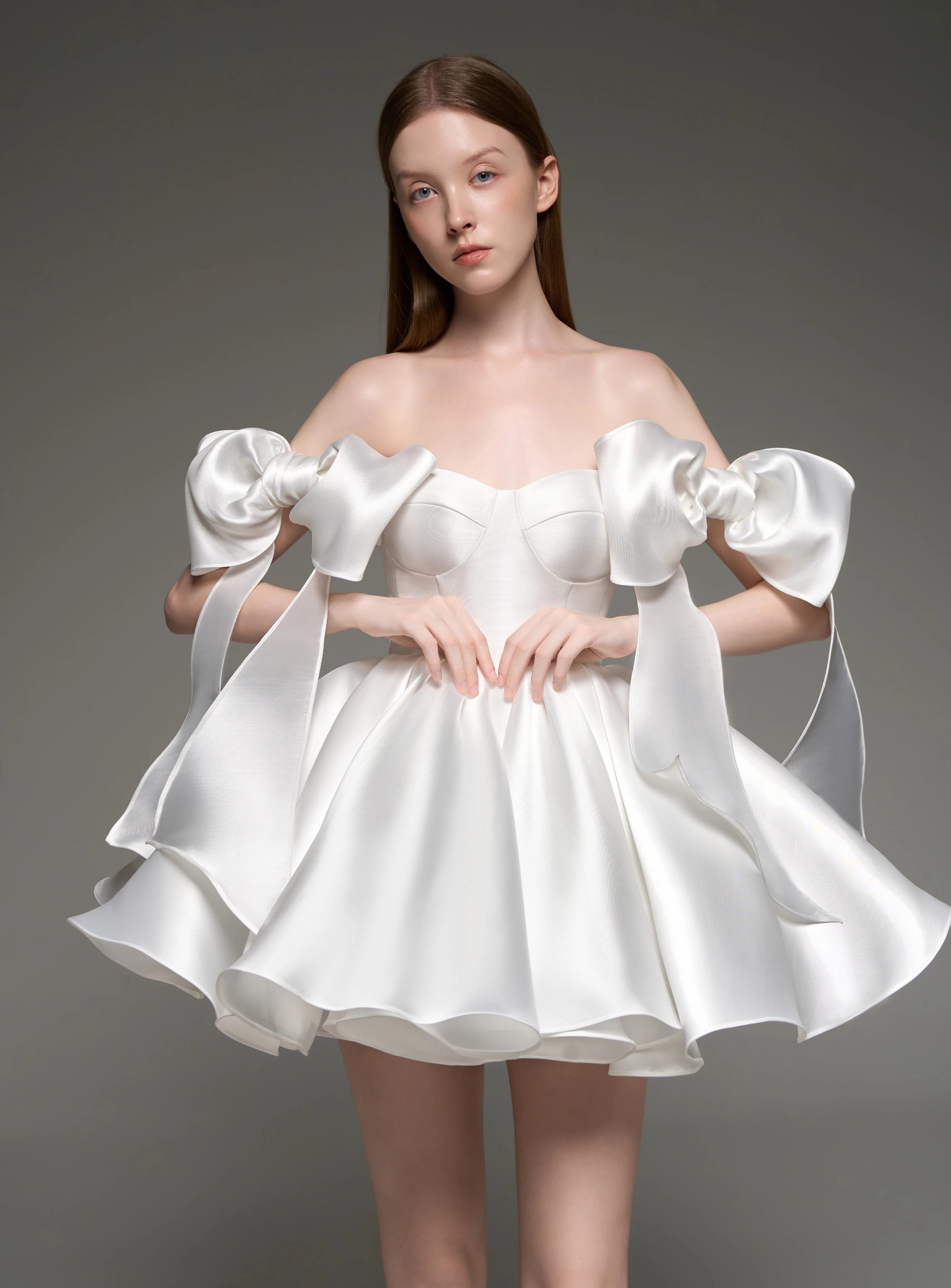 A model wearing a white satin off-the-shoulder mini dress with dramatic oversized bow details on the sleeves and a flared skirt, posed against a dark gray studio background.