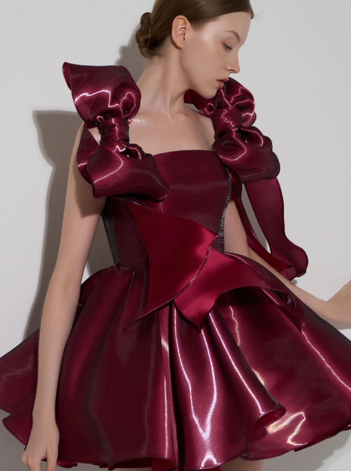 A model wearing a structured, floor-length dress made of shiny deep burgundy satin. The dress features dramatic oversized ruffle details on the shoulders, a fitted bodice, and a flared skirt, set against a plain light gray studio background.