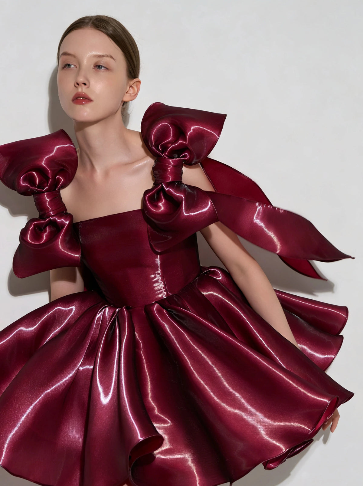 A model wearing a custom formal dress made of shiny deep burgundy satin, featuring oversized structured shoulder bows and a voluminous skirt, set against a plain light gray background.