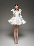 A model wearing a white custom formal dress with dramatic ruffled shoulder details and a flared skirt, standing against a dark gray studio background. The brand name 'Lusilk' is displayed at the top.