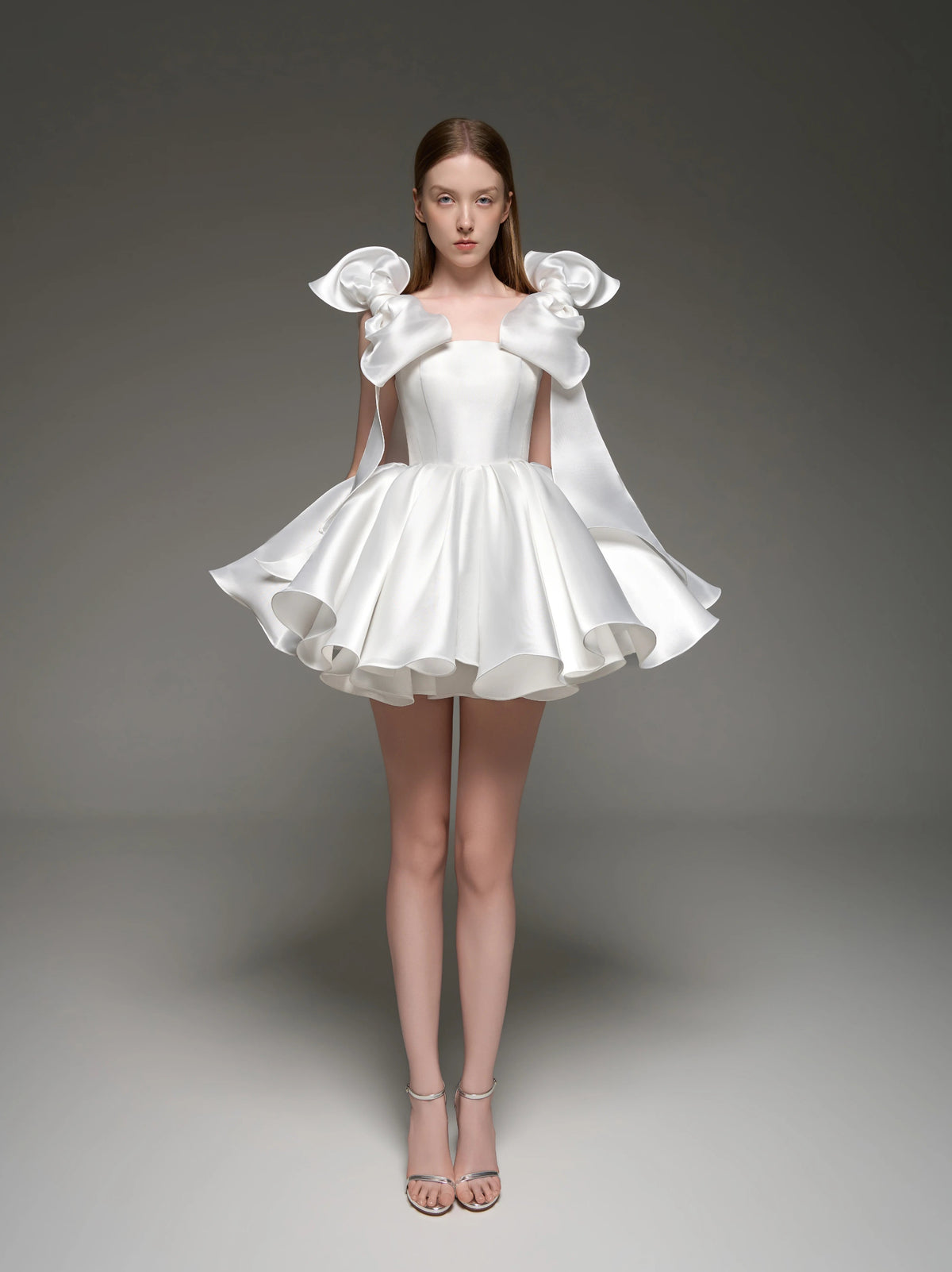 A model wearing a white custom formal dress with dramatic ruffled shoulder details and a flared skirt, standing against a dark gray studio background. The brand name 'Lusilk' is displayed at the top.