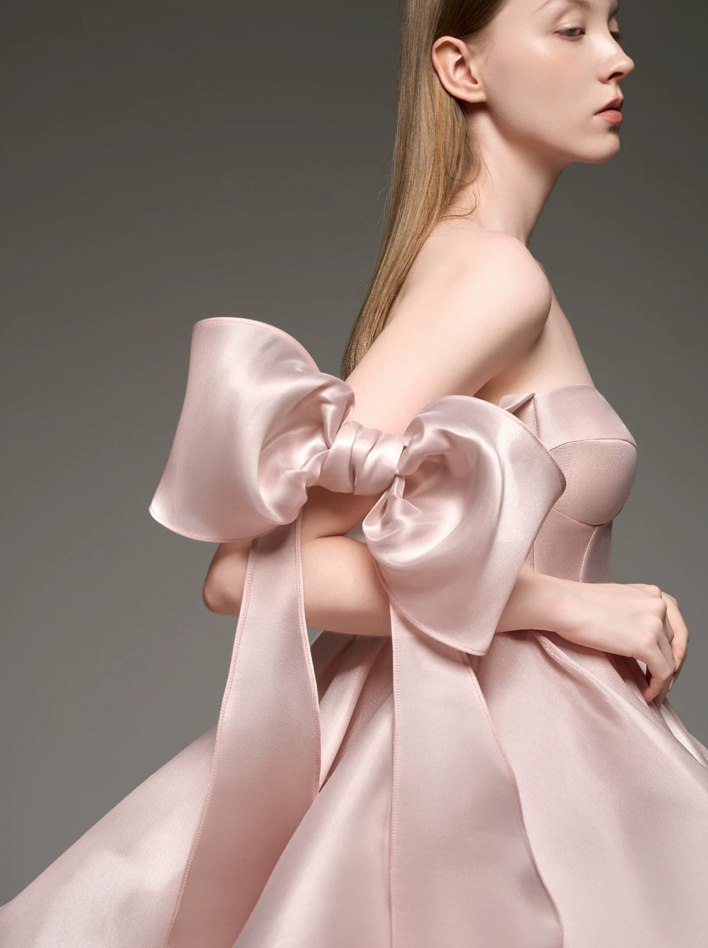 A model wearing a strapless formal dress made of shiny pale pink satin, featuring a massive oversized bow on one shoulder and a voluminous skirt, set against a dark gray studio background.