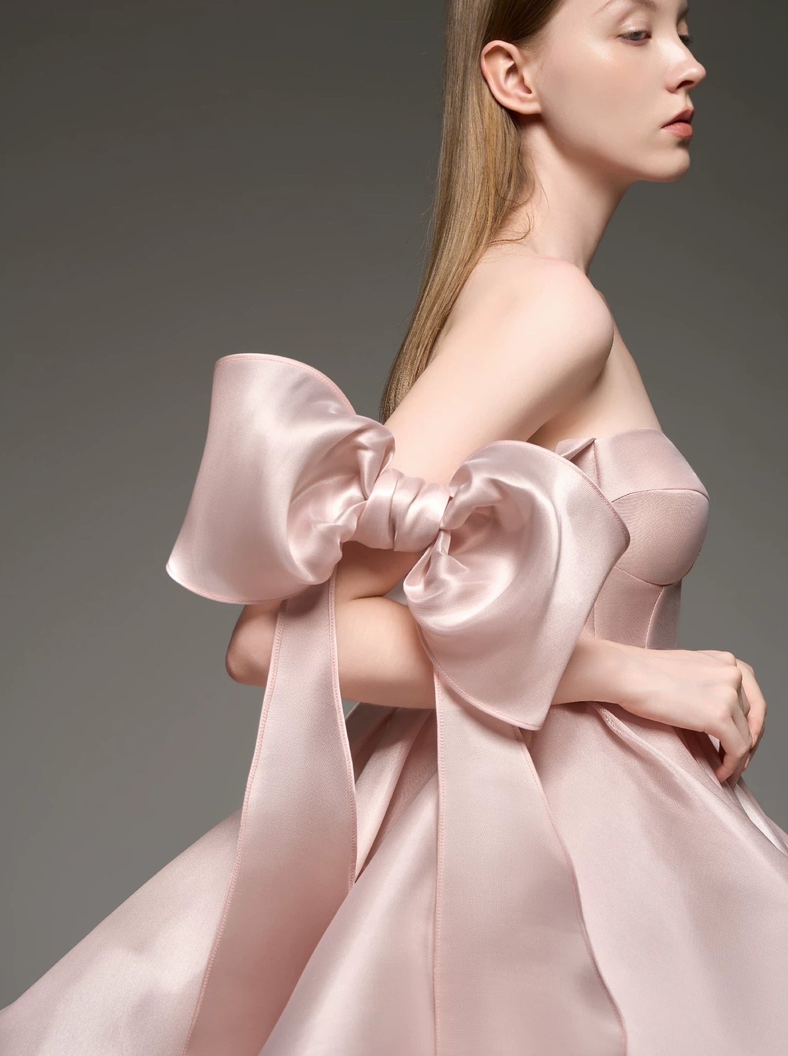 A model wearing a strapless formal dress made of shiny pale pink satin, featuring a massive oversized bow on one shoulder and a voluminous skirt, set against a dark gray studio background.