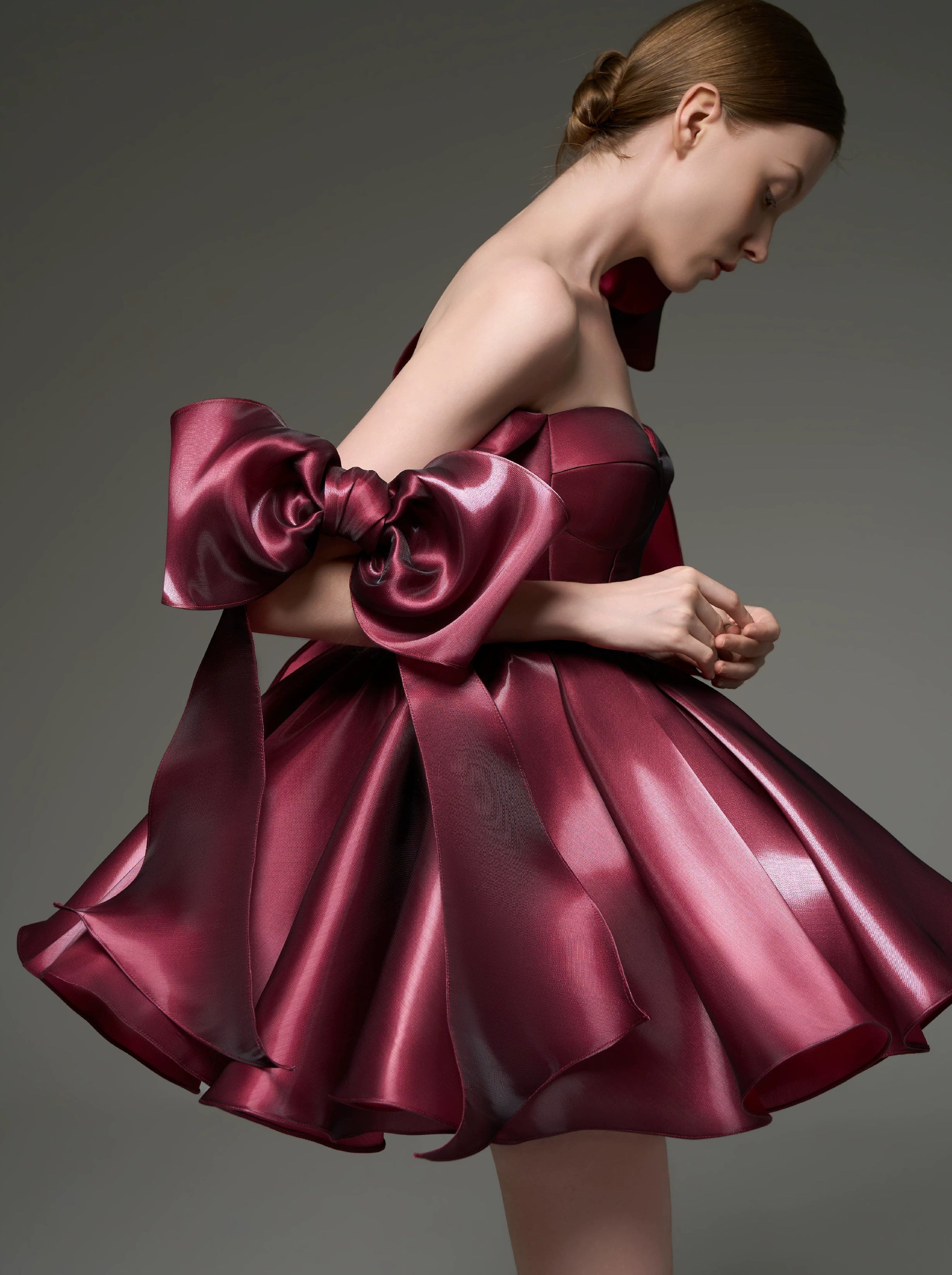 A model wearing a strapless formal dress made of shiny burgundy satin, featuring a large oversized bow on one shoulder and a flared skirt, posed against a dark gray studio background.