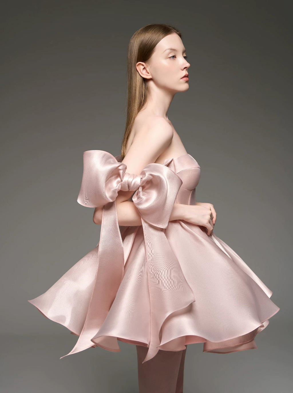 A model wearing a light pink strapless dress with oversized bow details on the shoulders and a flared skirt, posed against a dark gray studio background.
