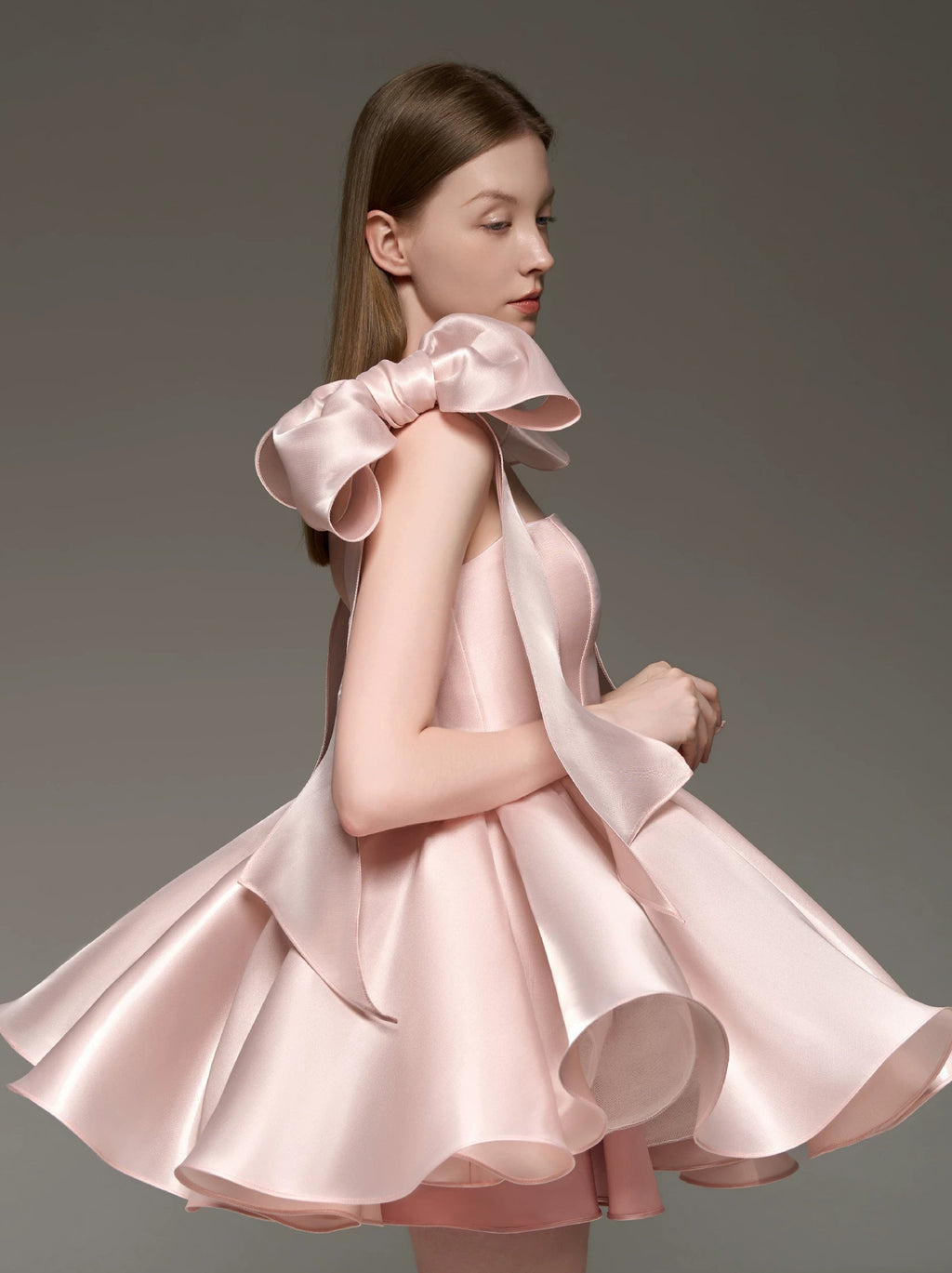 A woman modeling a light pink satin dress with a dramatic oversized bow at the shoulder and a flared skirt, photographed against a plain gray studio background.
