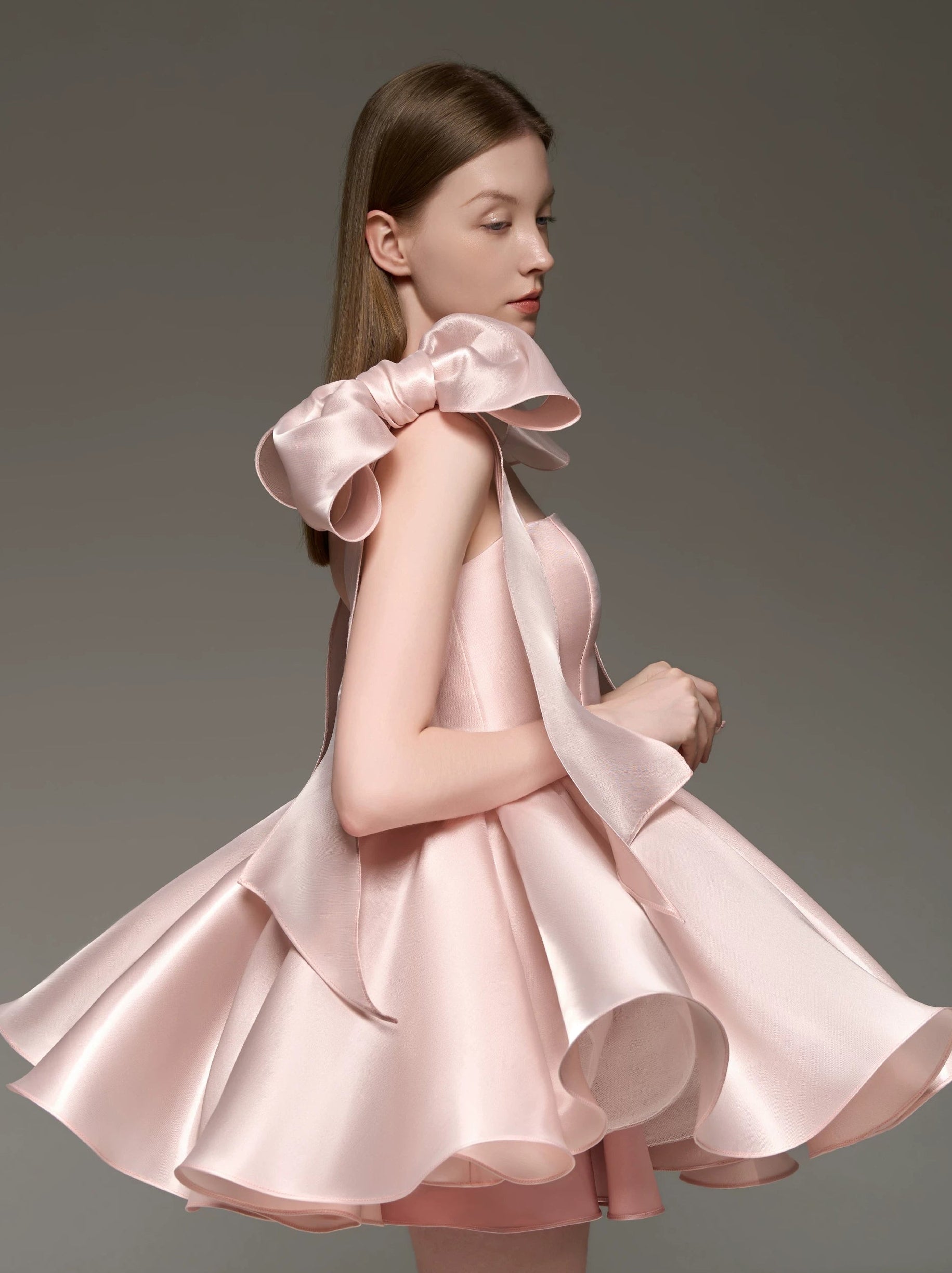 A woman modeling a light pink satin dress with a dramatic oversized bow at the shoulder and a flared skirt, photographed against a plain gray studio background.
