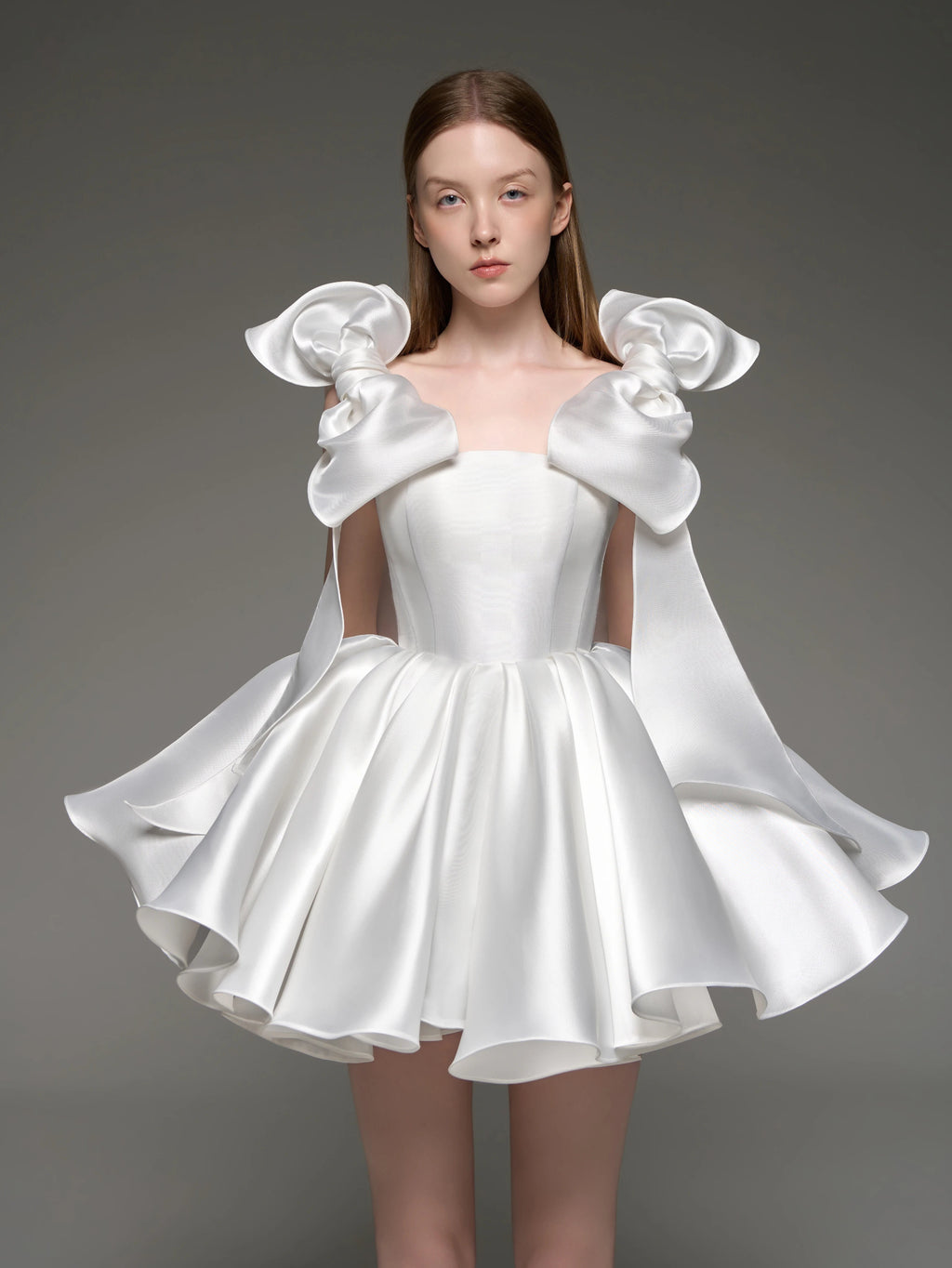 A model wearing a white satin formal dress with dramatic oversized shoulder bows and a flared skirt, photographed against a dark gray studio background.