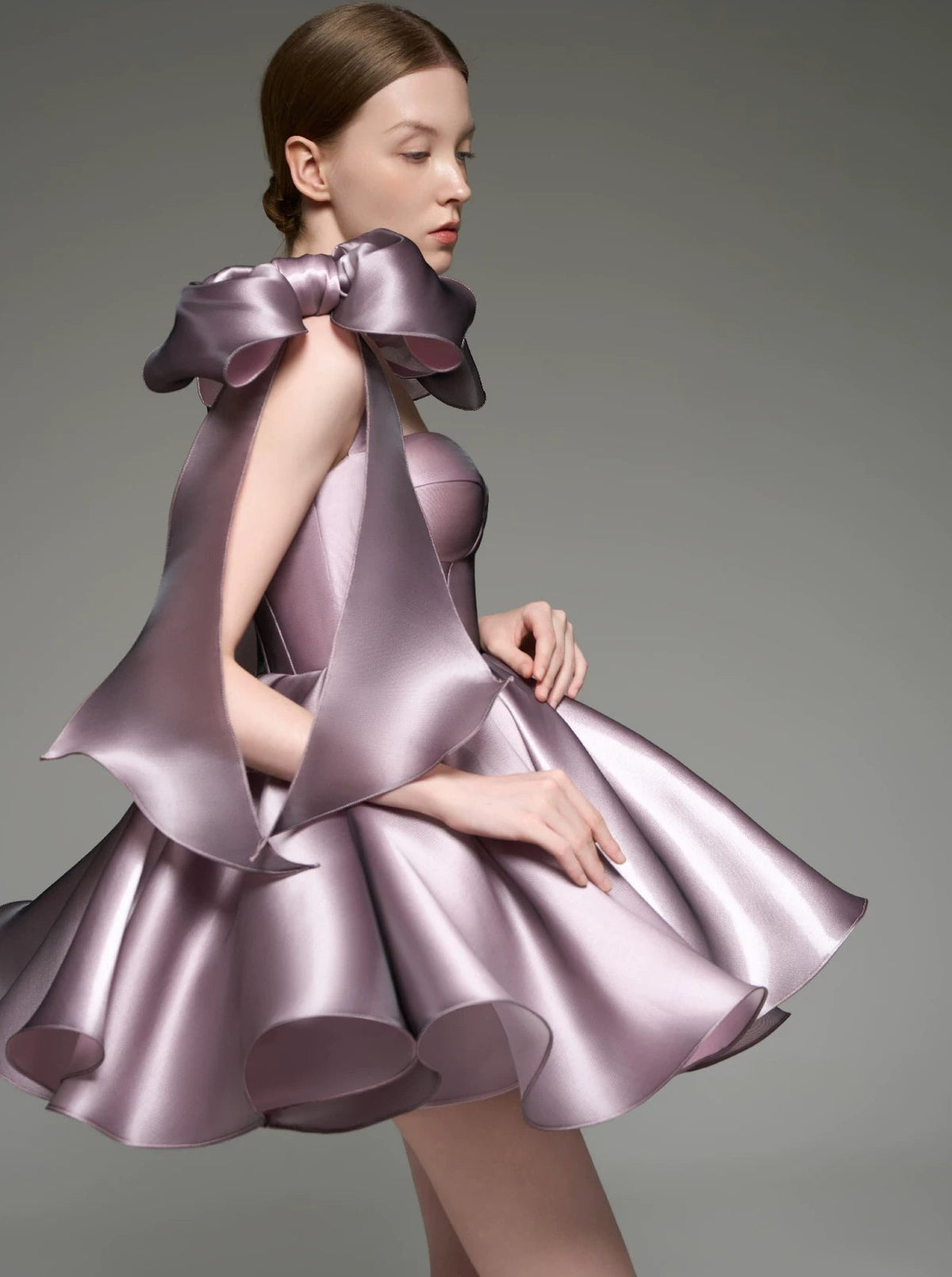 A model wearing a custom formal dress made of shiny lavender-purple satin, featuring a structured bodice, oversized bow-like shoulder details, and a flared skirt, set against a plain gray studio background.