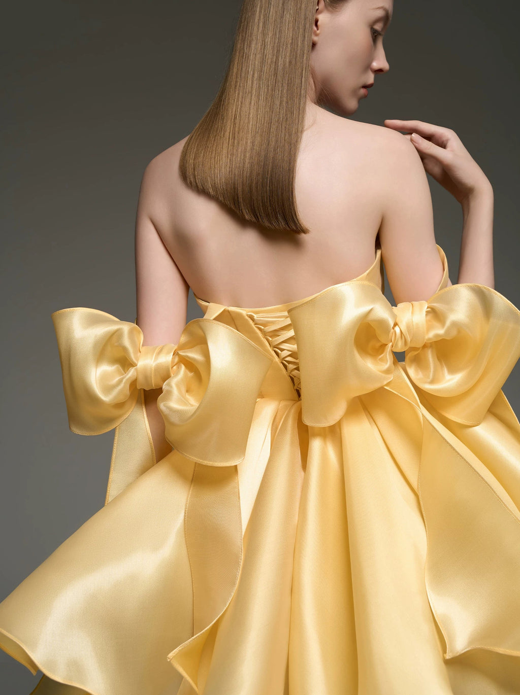 A woman shown from the back wearing a strapless formal dress made of shiny pale yellow satin, with oversized bow details at the shoulders and a large bow across the back. The photo is set against a dark gray studio background.